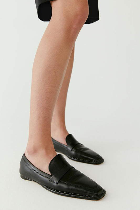 Side view showing the loafers worn with a black skirt. The low heel and minimal seam lines emphasize the refined, understated design.