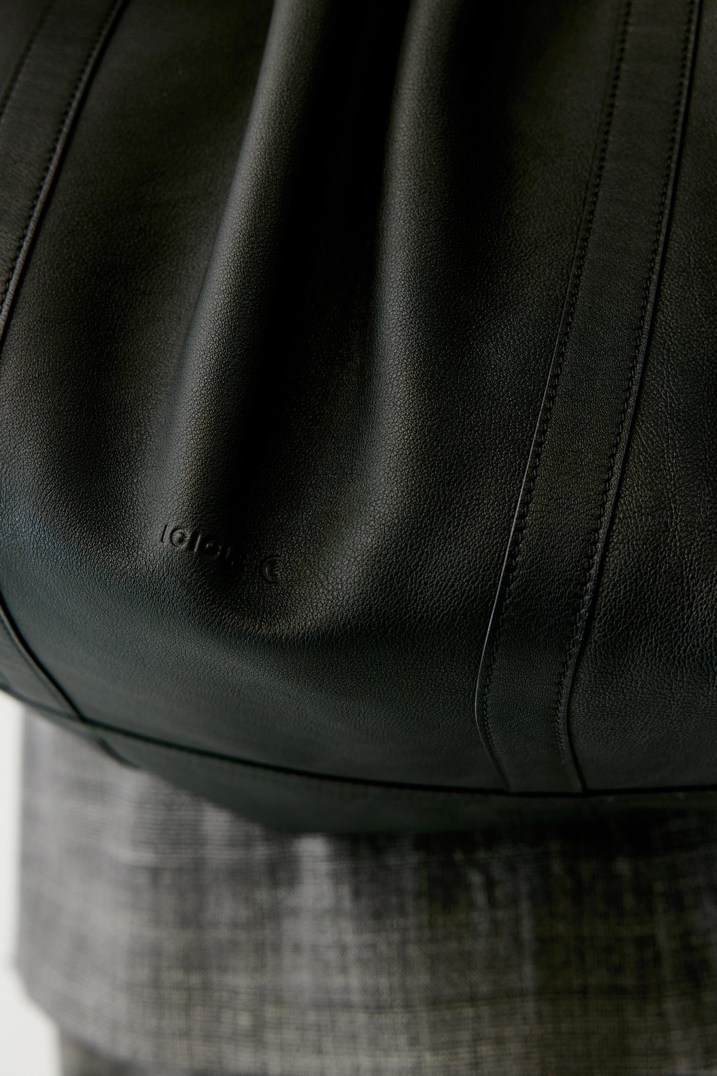 Close-up of a black leather shoulder bag with visible grain texture and a subtle embossed logo, worn over gray plaid fabric.
