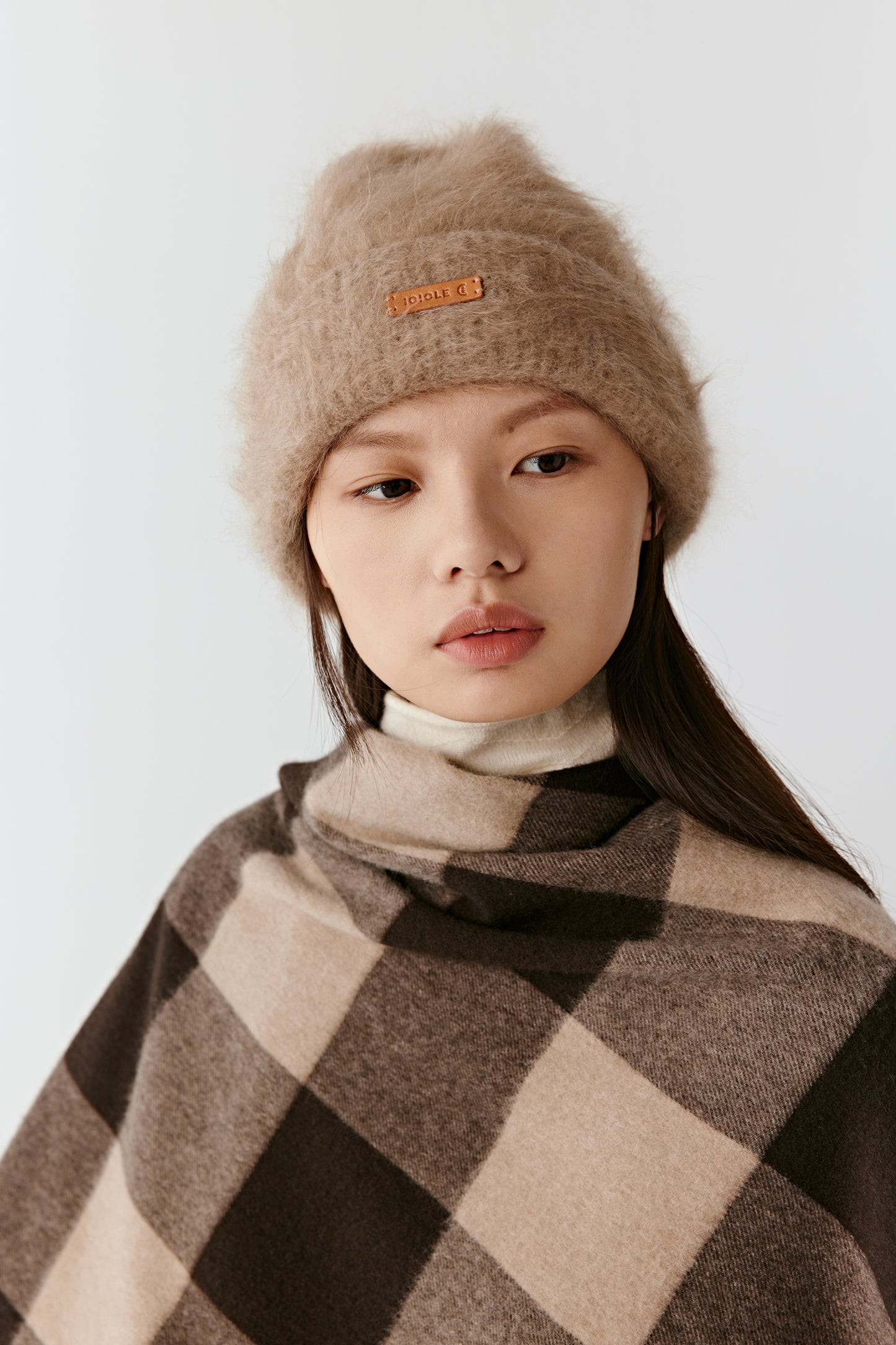 Woman wearing a taupe knit beanie and a brown checkered poncho, looking forward against a light background.