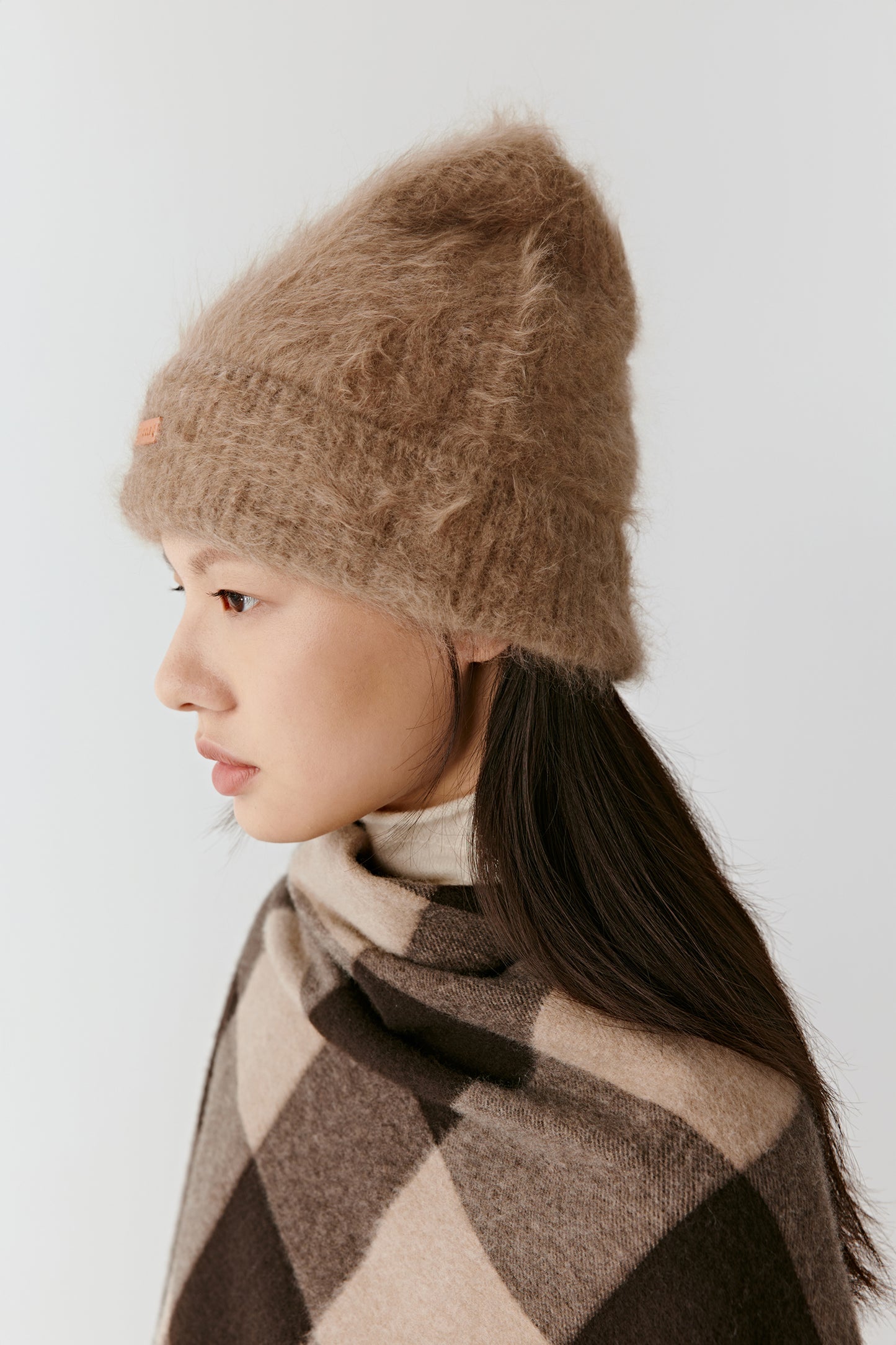Side view of a woman wearing a taupe knit beanie and a brown checkered poncho, against a light background.