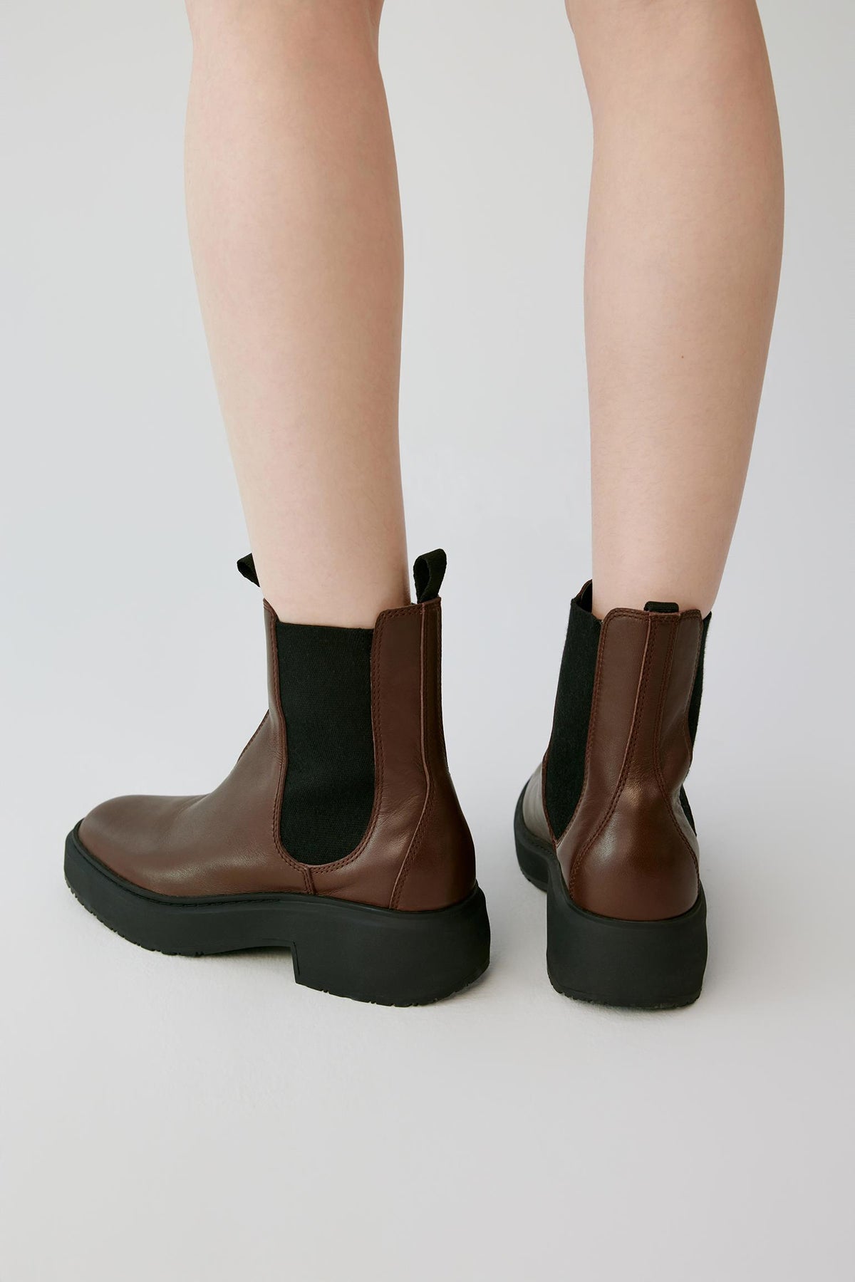 Brown and black Chelsea-style ankle boots shown from the back on a model's feet, against a clean light gray studio background.