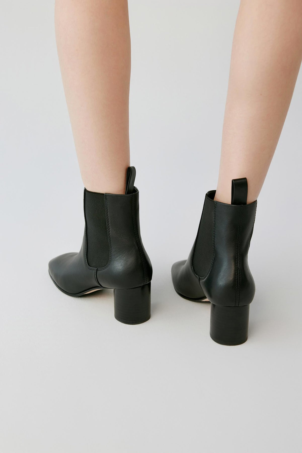 Black leather ankle boots with block heels, shown from the back on a model's feet against a light gray studio background.