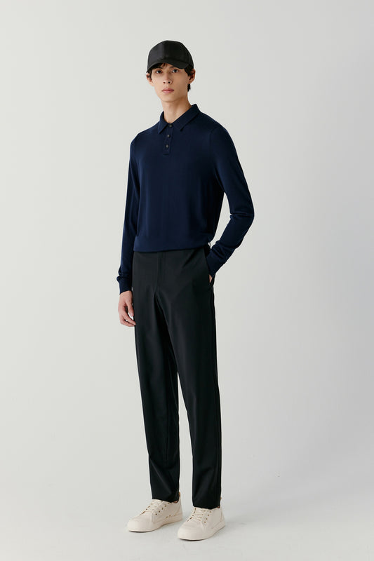 A man stands in a navy merino wool polo and black trousers, facing forward in a relaxed posture.