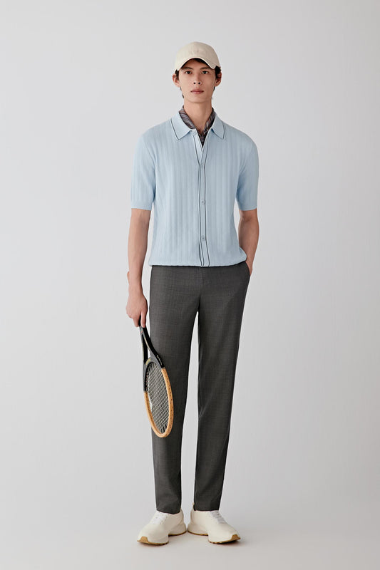 Man wearing a light blue knitted cotton short-sleeve shirt with relaxed straight fit, styled with dark grey trousers and beige cap, full-length front view.