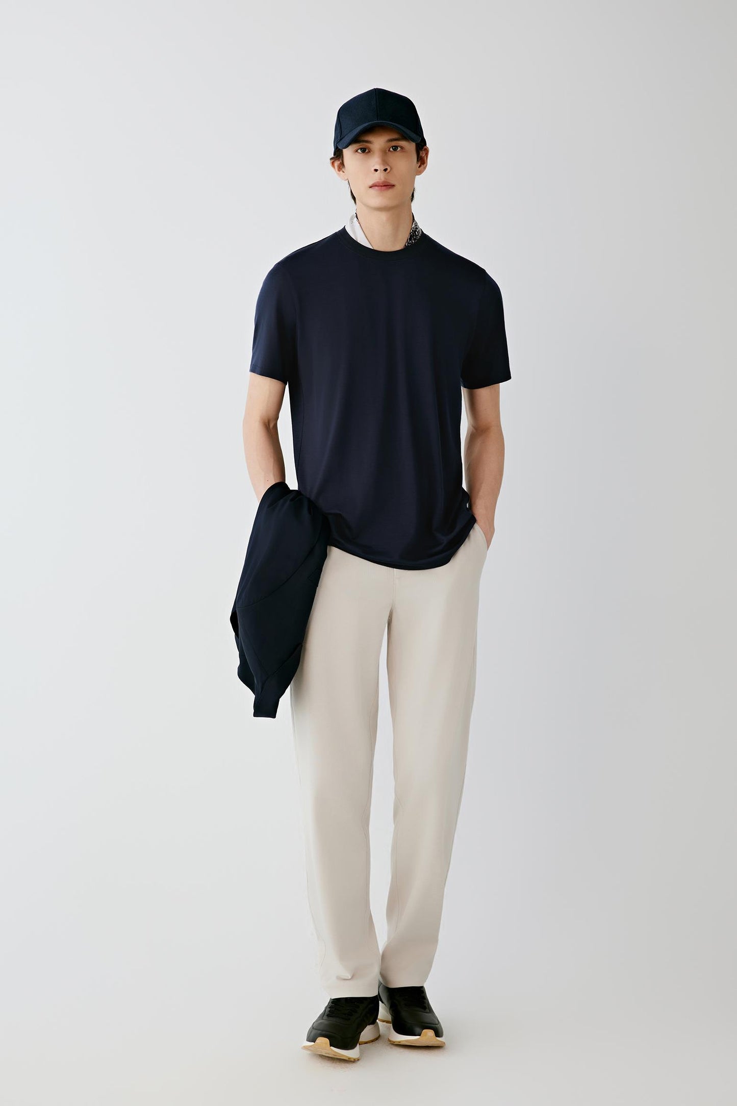 A man stands against a light gray studio background wearing a dark navy short-sleeve crewneck t-shirt, off-white trousers, black sneakers, and a dark navy baseball cap. He is holding a matching navy jacket over his arm.