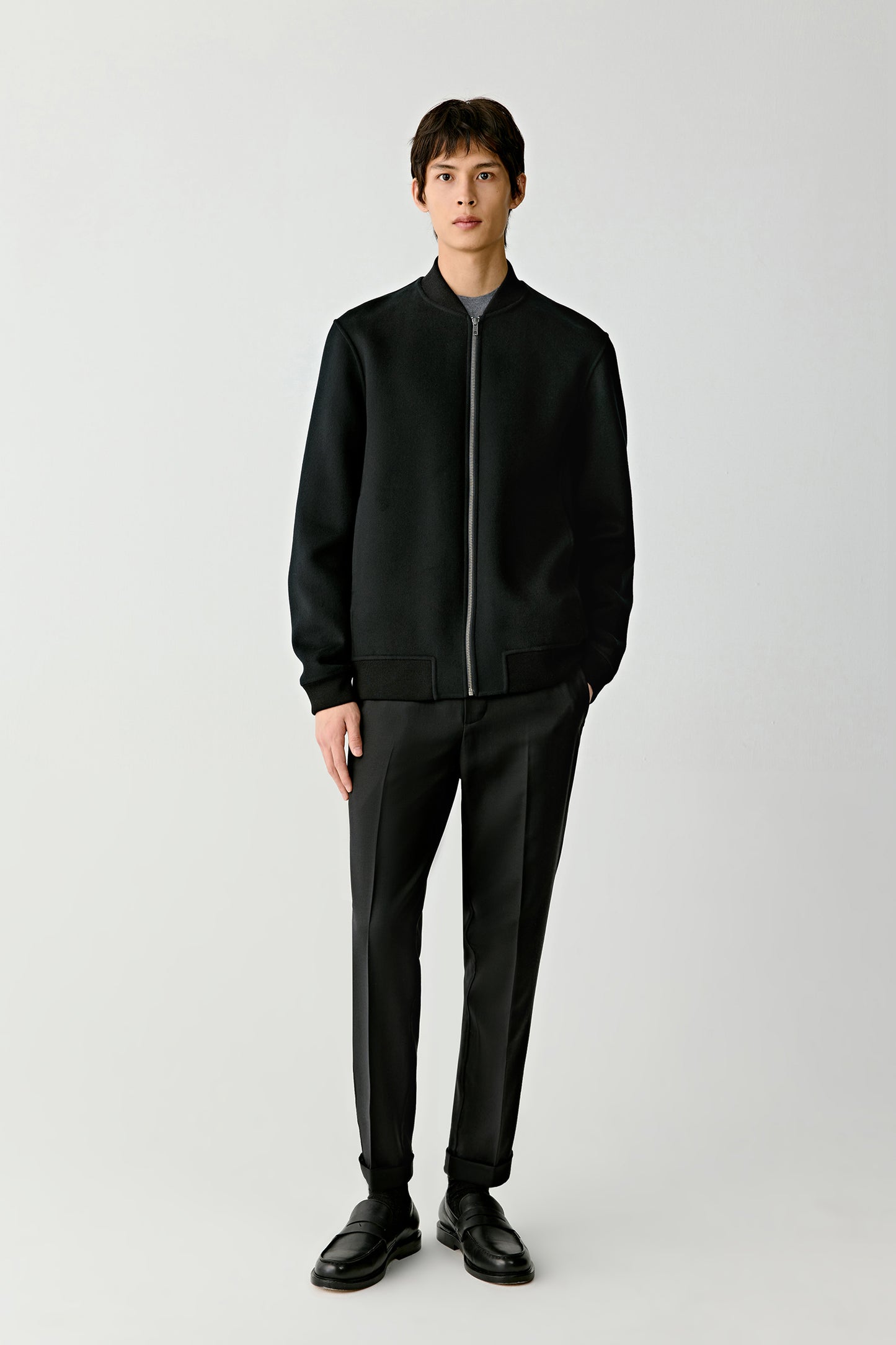 Front full-length view of a man wearing a deep black double-faced wool jacket with a minimalist silhouette and tailored trousers.