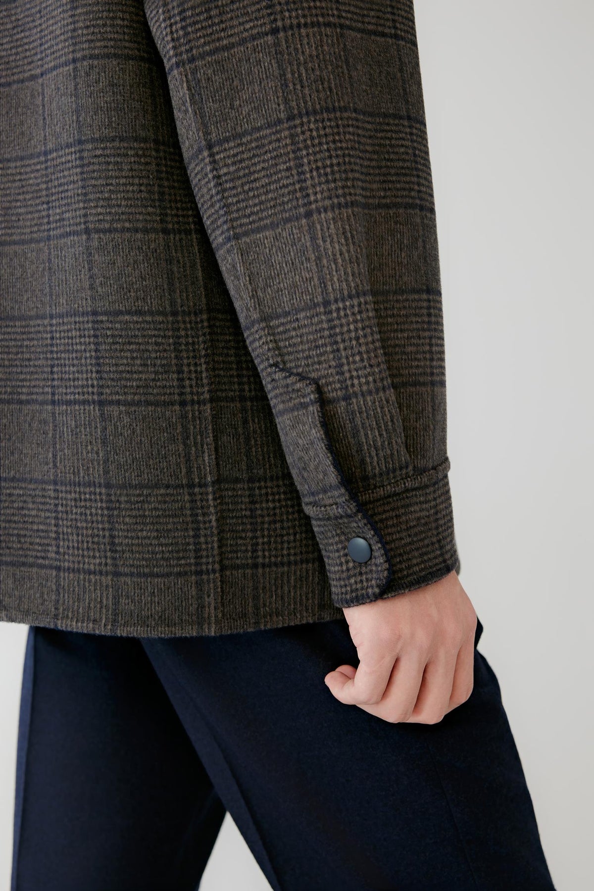 Close-up of a person wearing a dark brown and charcoal plaid wool-blend jacket with a buttoned cuff, paired with navy trousers, against a light gray background.