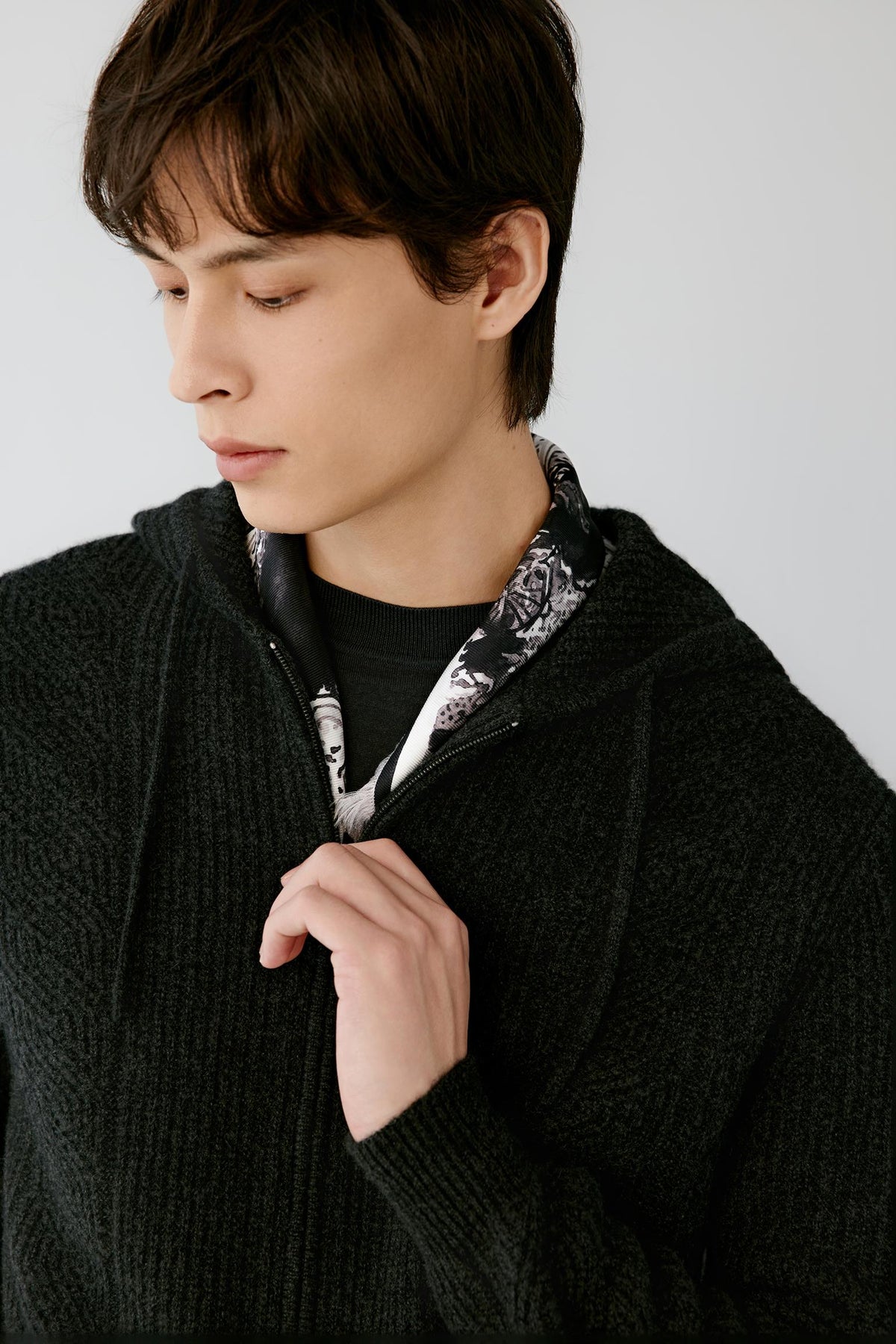 A close-up of a man wearing a black ribbed knit zip-up hoodie over a black shirt, with a black, white, and gray patterned neck scarf visible at the collar, set against a plain light gray background.
