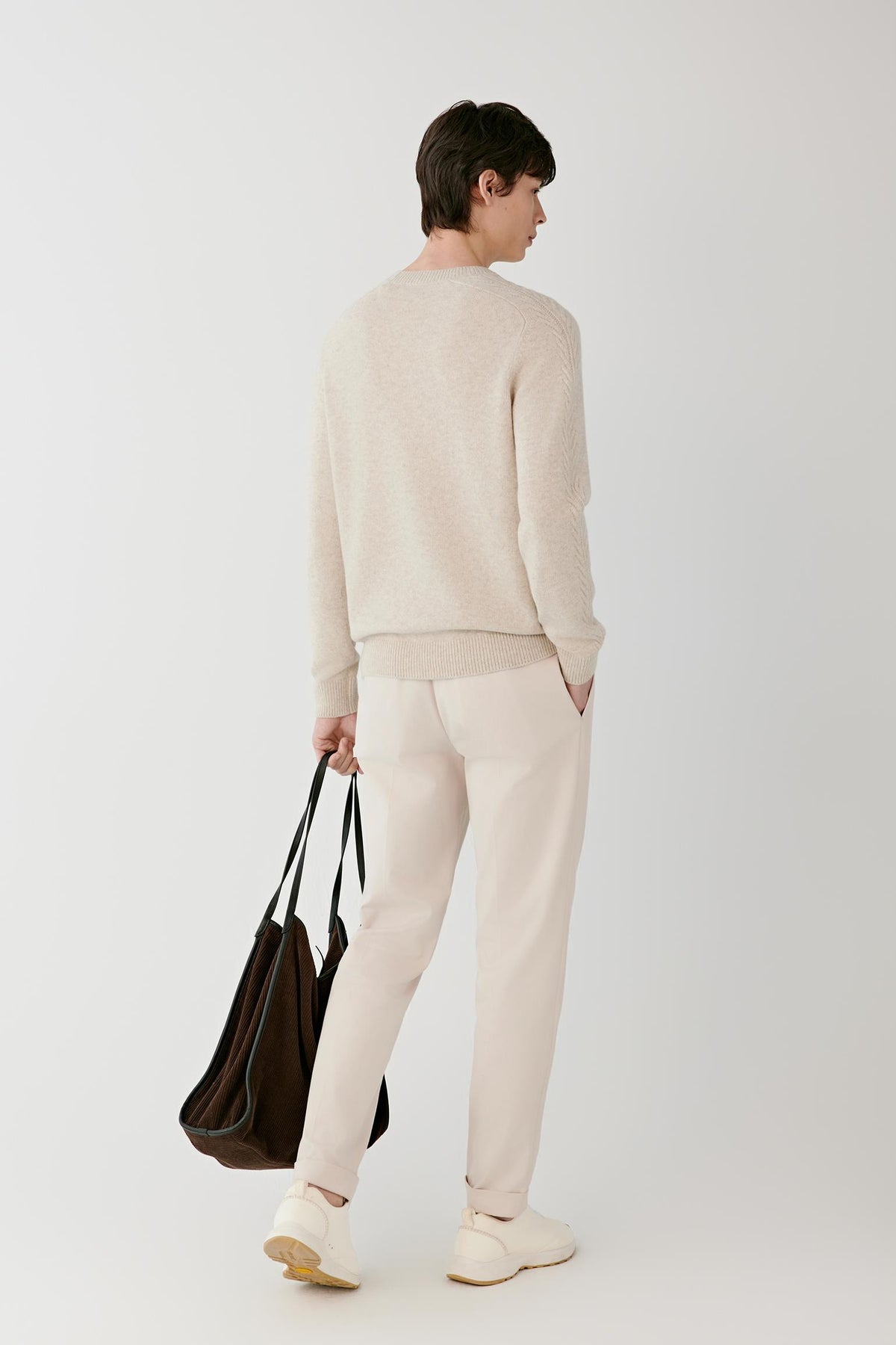 A man is shown from behind wearing a light beige knit sweater and matching trousers, holding a dark brown leather tote bag against a plain light gray studio background.