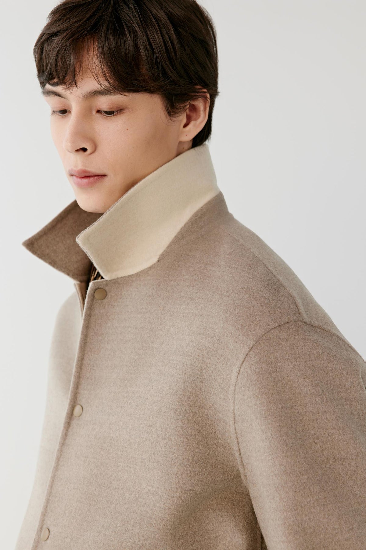 A close-up side profile of a man wearing a beige wool-blend coat with a contrasting cream-colored collar, set against a plain light gray studio background.