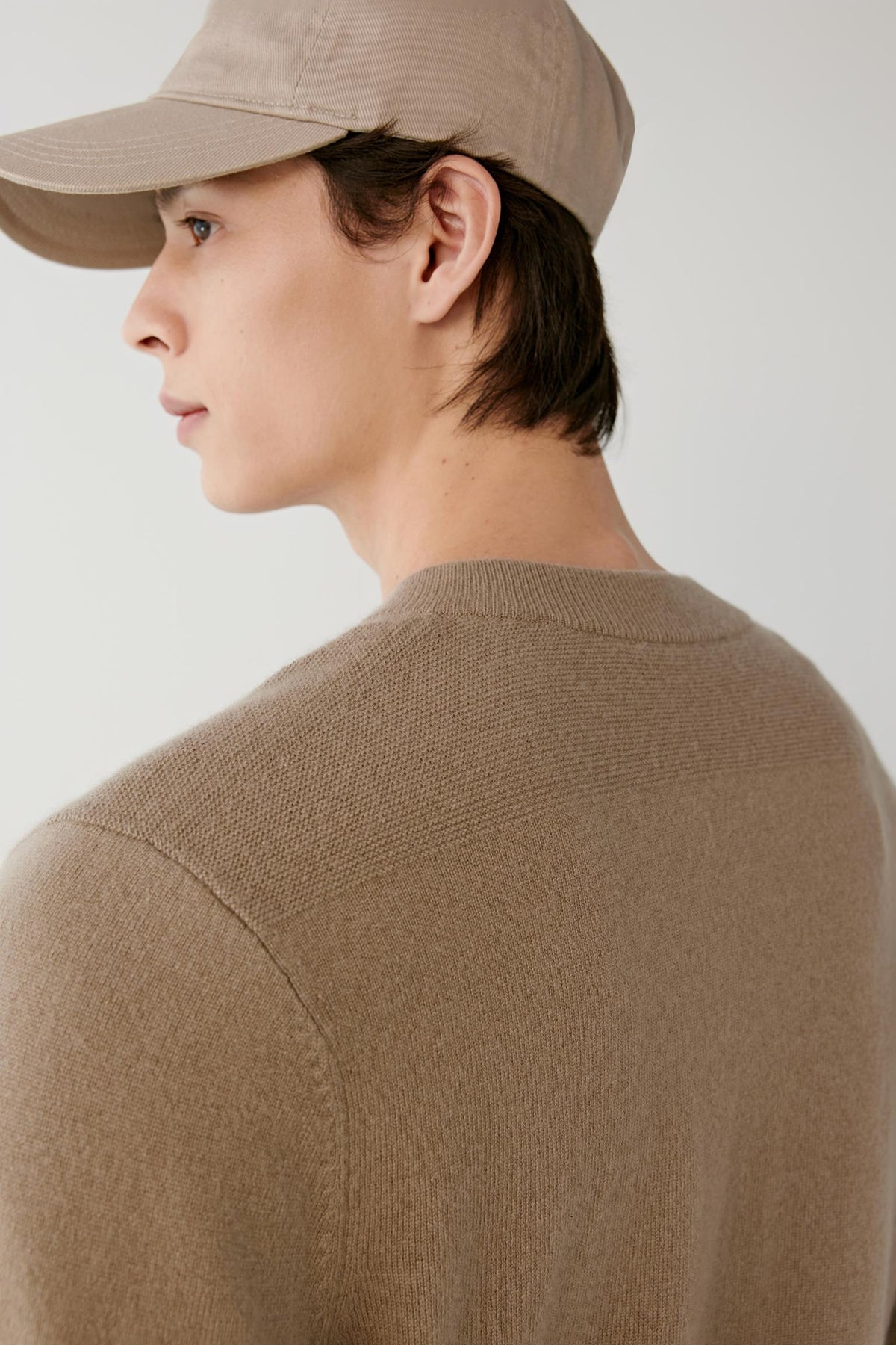 A man shown in profile wearing a beige knit sweater and a matching beige baseball cap against a plain light gray background.