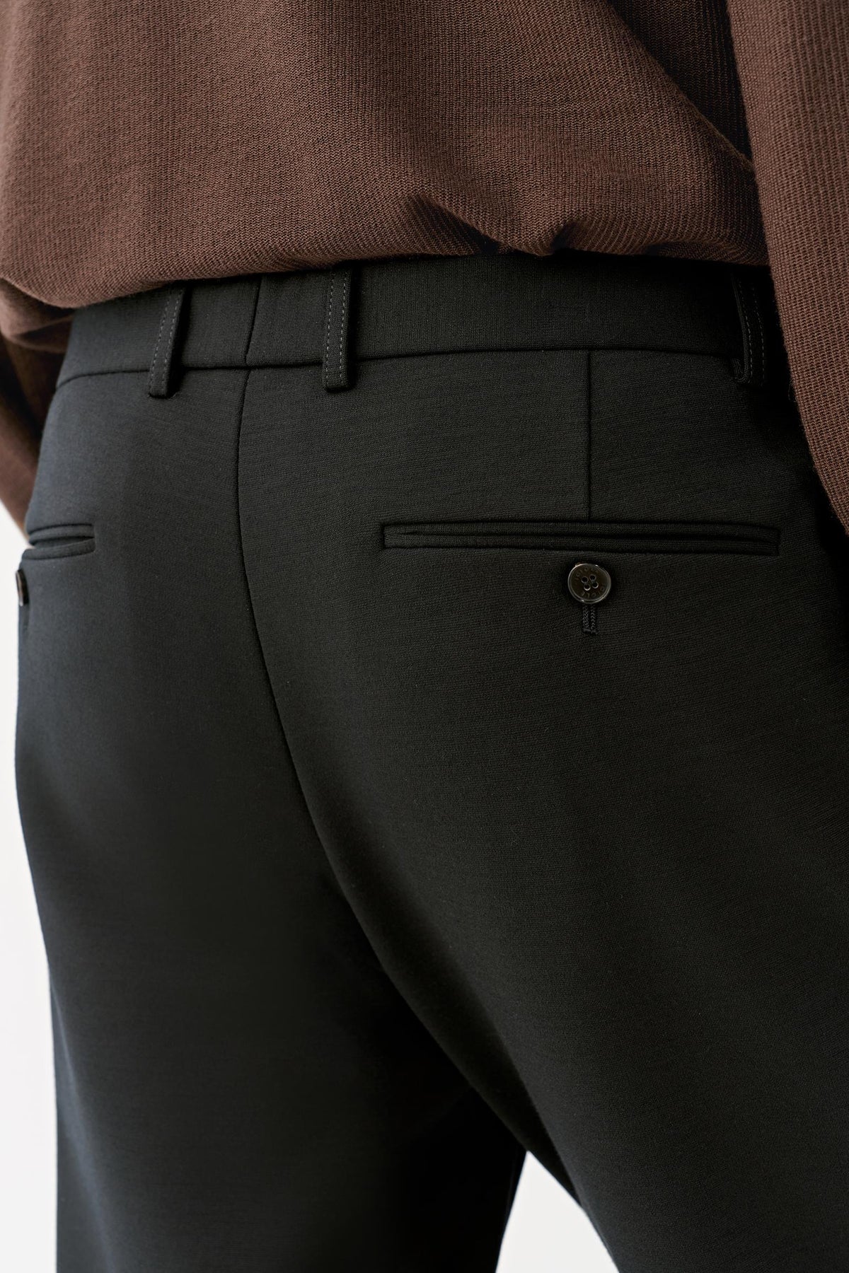 Back detail highlighting the waistband and buttoned welt pockets of the black wool trousers.