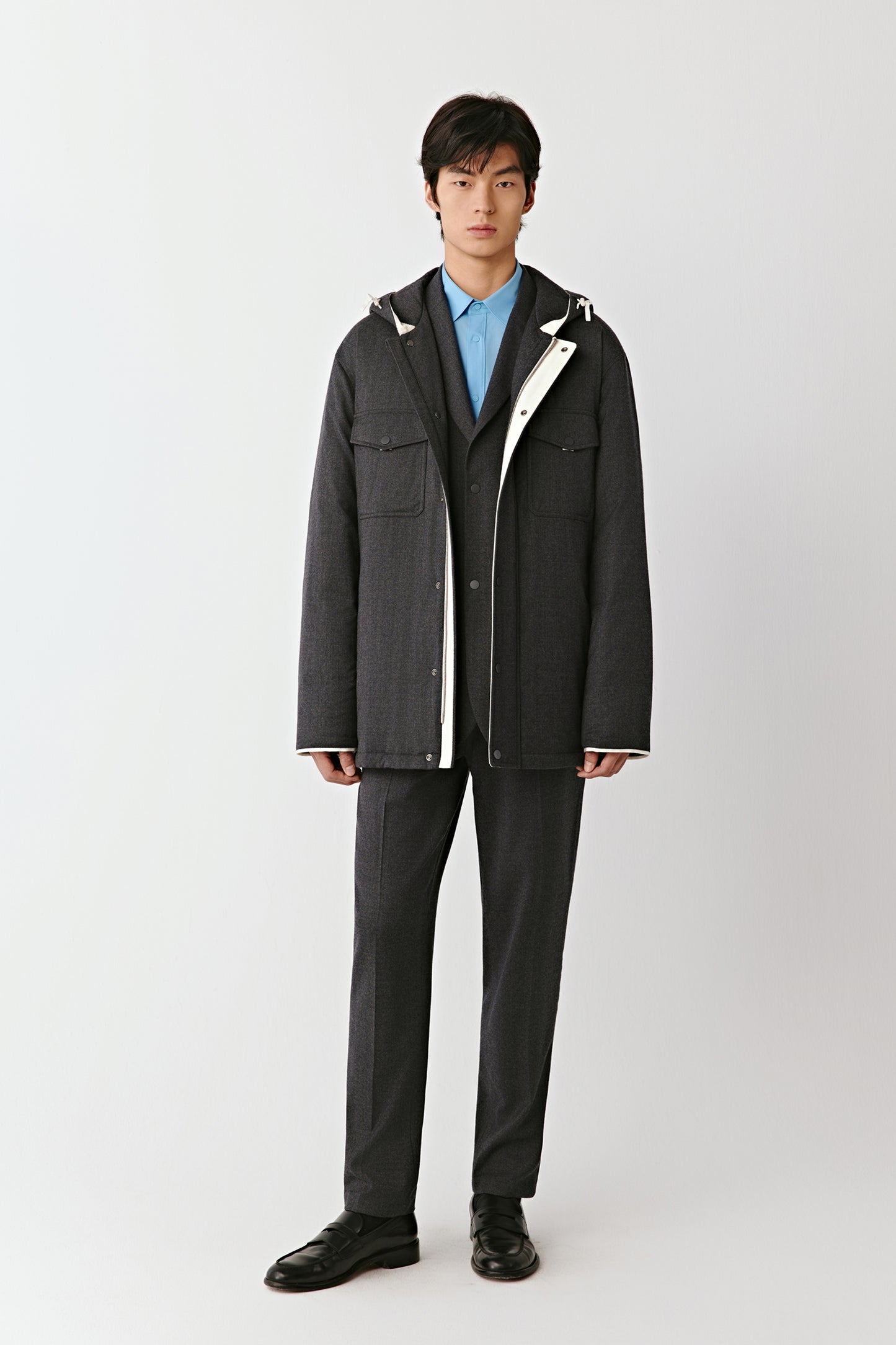 Full-length front view of a man wearing a grey wool flannel quilted jacket layered over a blue shirt and black trousers.