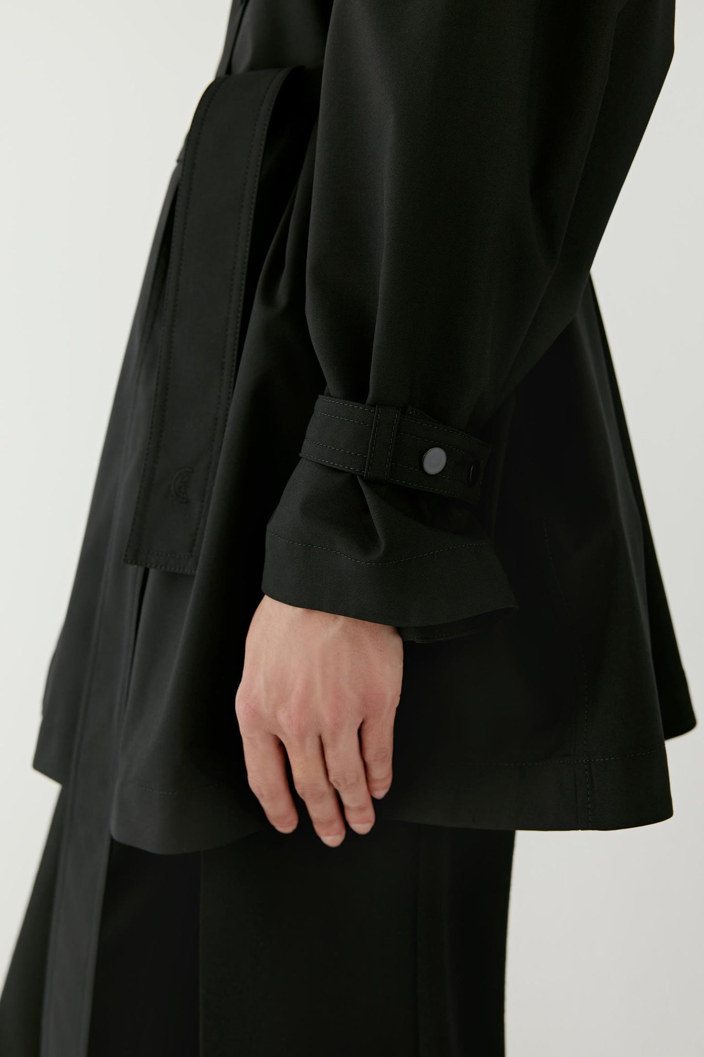 Close-up detail of the sleeve and side seam of the black wool and silk trench coat, showing the smooth matte texture of the fabric and the clean tailored stitching.