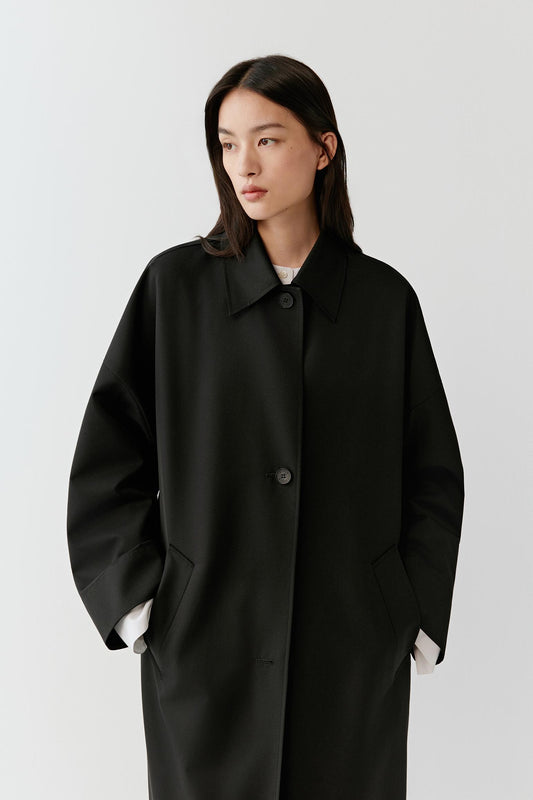 Three-quarter view of the black wool trench coat showing the relaxed shoulders, straight cut and fluid drape.
