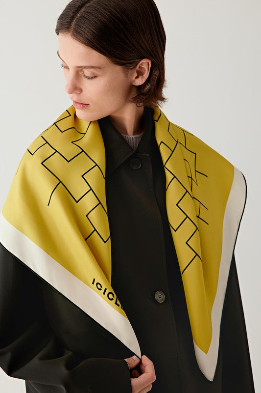 Three-quarter view showing the scarf draped around the neck and shoulders over a black garment. The geometric motif unfolds across the yellow surface as the fabric falls naturally.