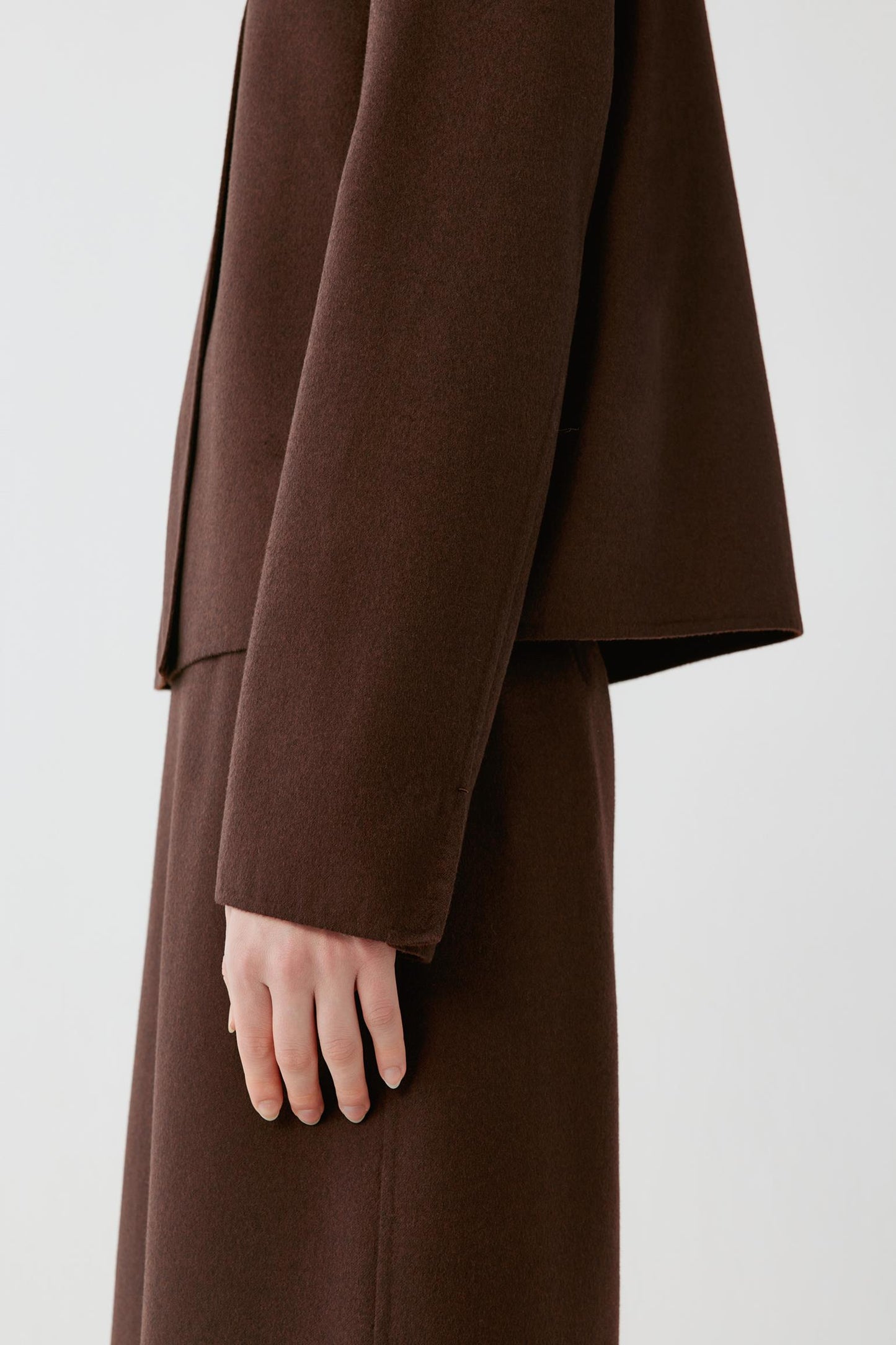 A close-up side view of a person wearing a dark brown wool coat with a matching long skirt or dress, showing the sleeve, hand, and drape of the fabric against a plain light gray background.