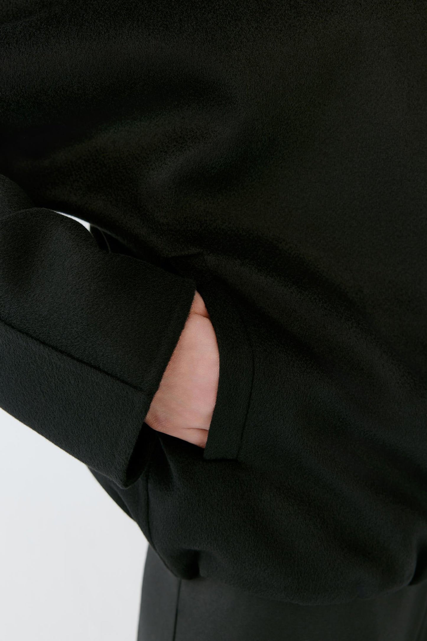 Close-up of a black coat sleeve with a hand tucked into the pocket, showing the fabric texture and cuff detail.