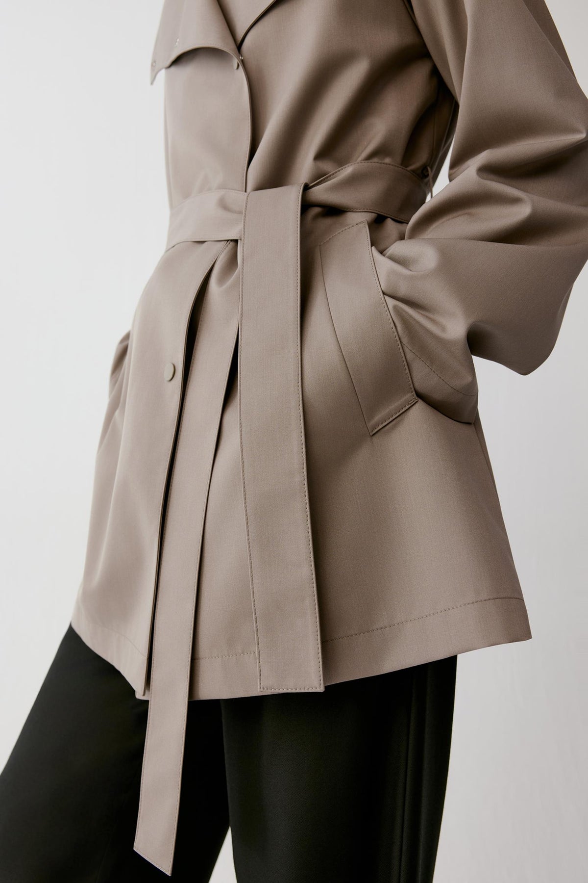 A close-up of a model wearing a taupe-brown belted trench coat with black pants, shown from the chest down against a light gray background.