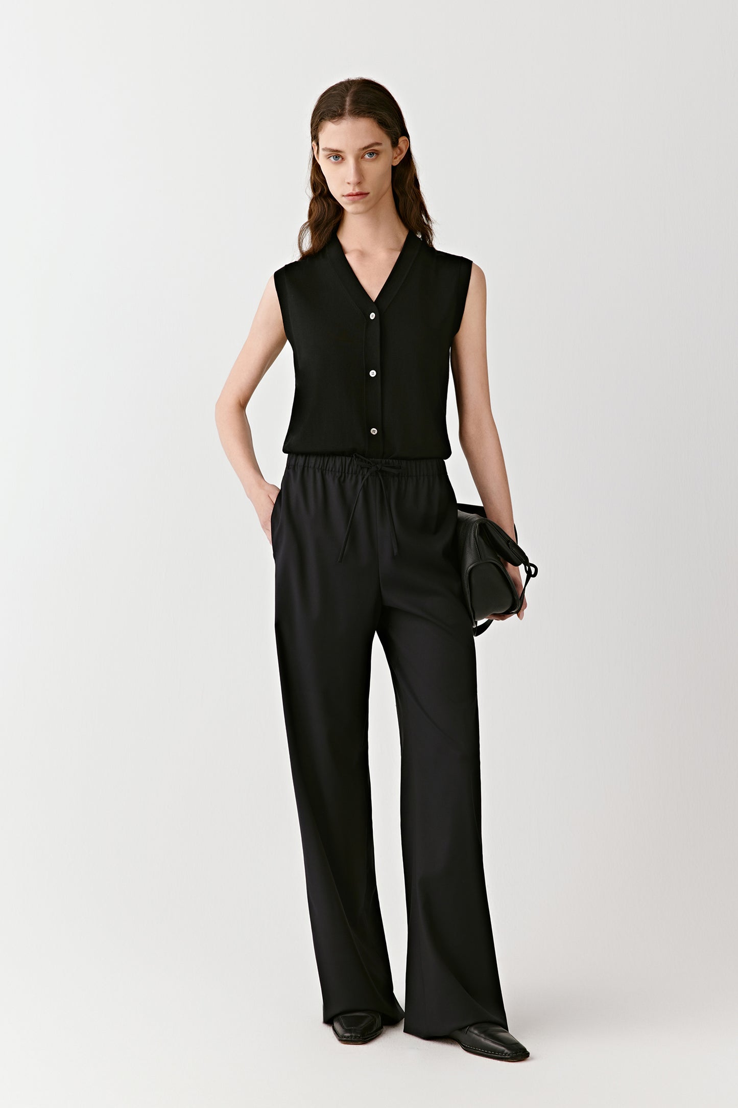 A woman stands in black wide-leg worsted wool trousers and a sleeveless top, softly lit against a neutral background.