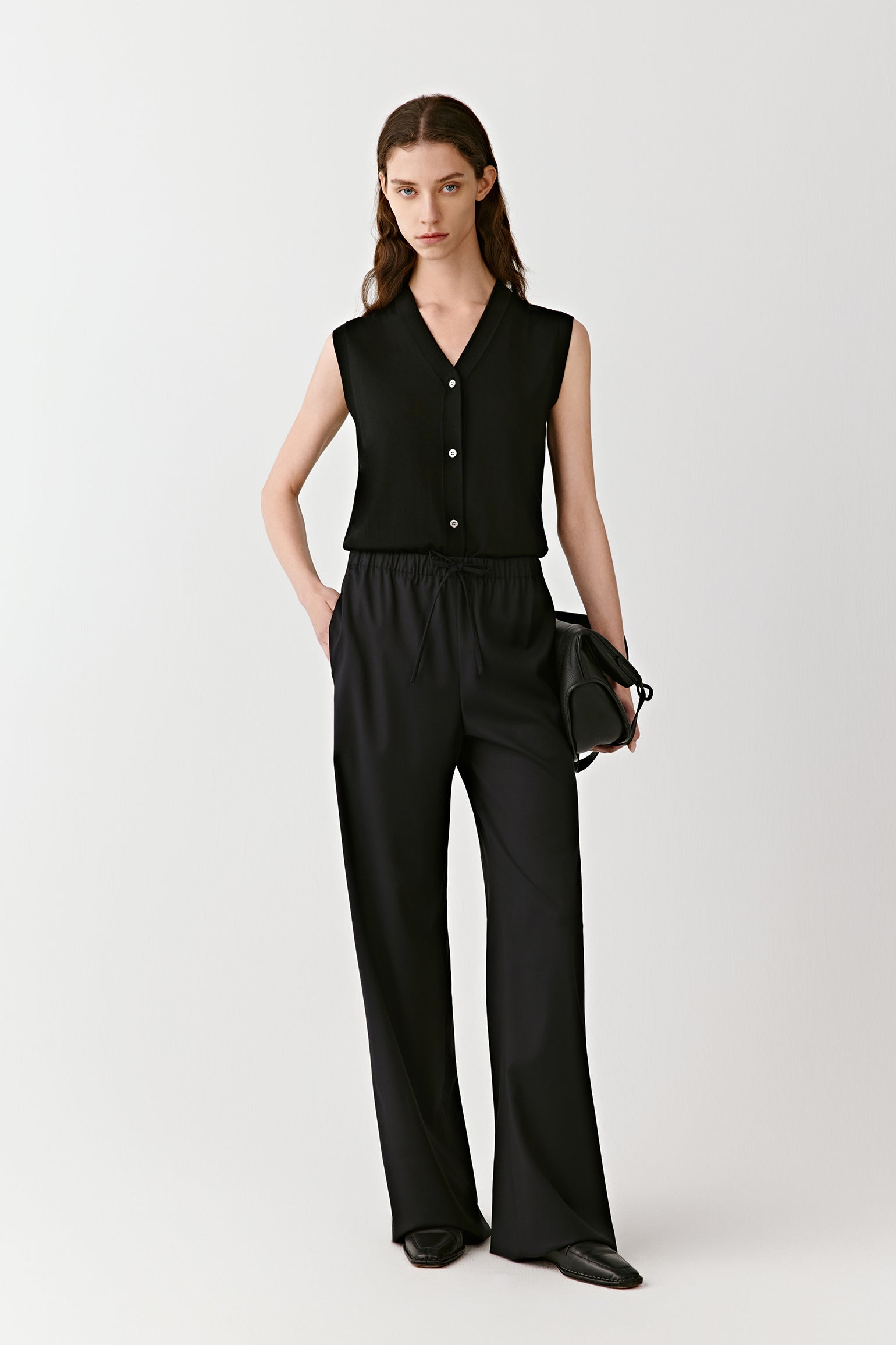 A woman stands in black wide-leg worsted wool trousers and a sleeveless top, softly lit against a neutral background.