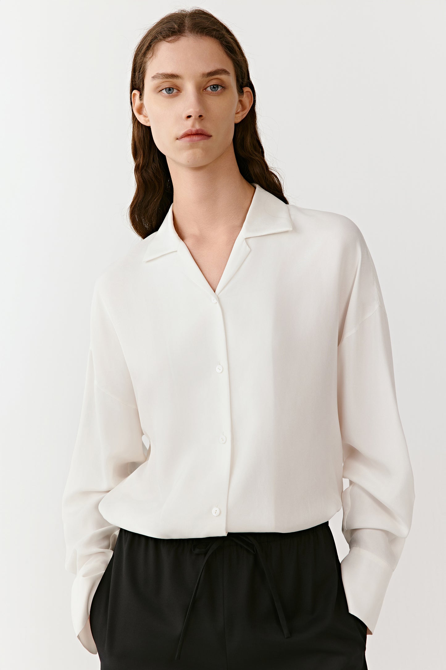 A woman stands in an ivory silk twill shirt and black trousers, the soft drape enhanced by gentle light.