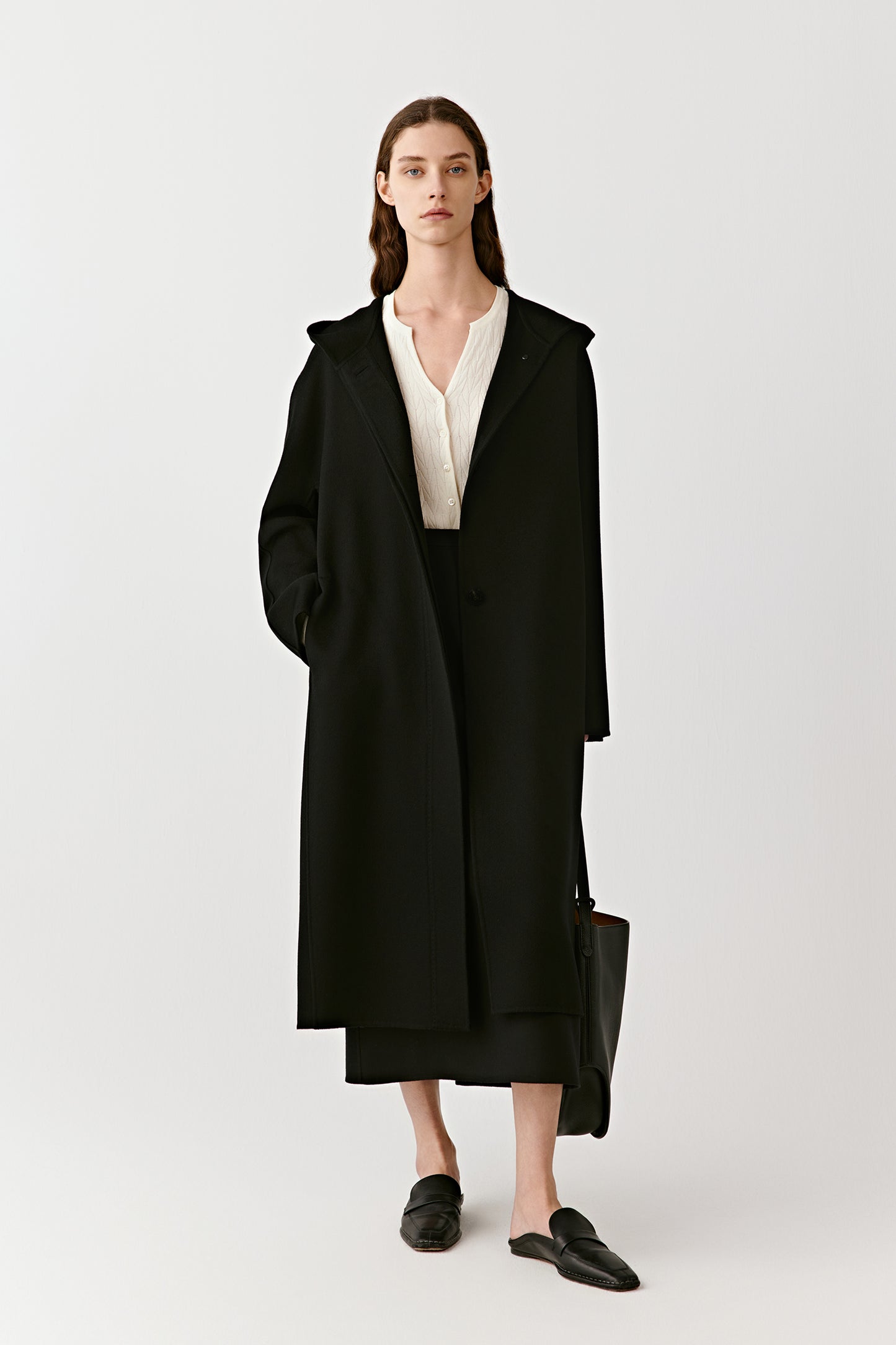 A woman stands in a long black hooded coat in double-faced cashmere, styled with minimalist shoes.