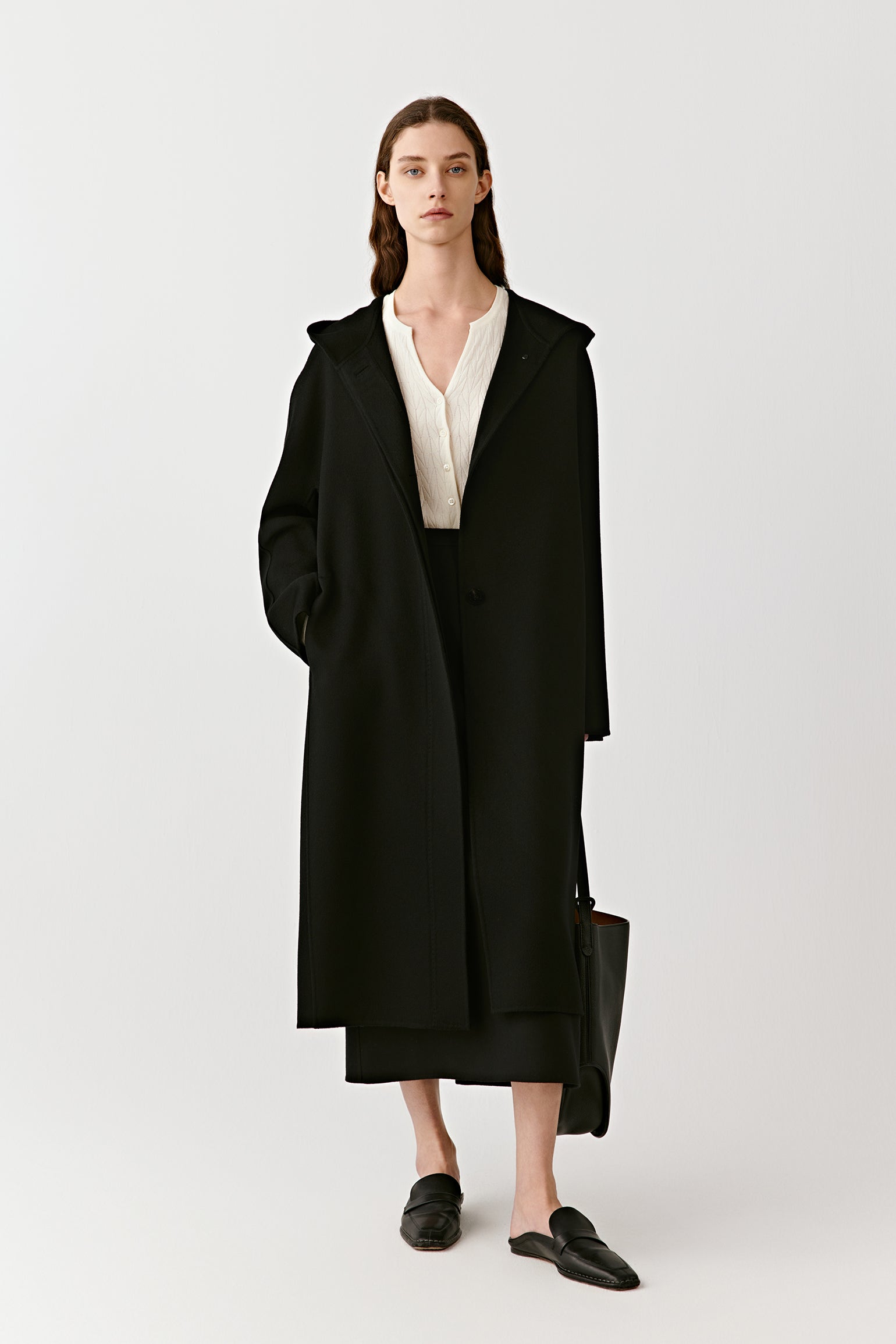 A woman stands in a long black hooded coat in double-faced cashmere, styled with minimalist shoes.