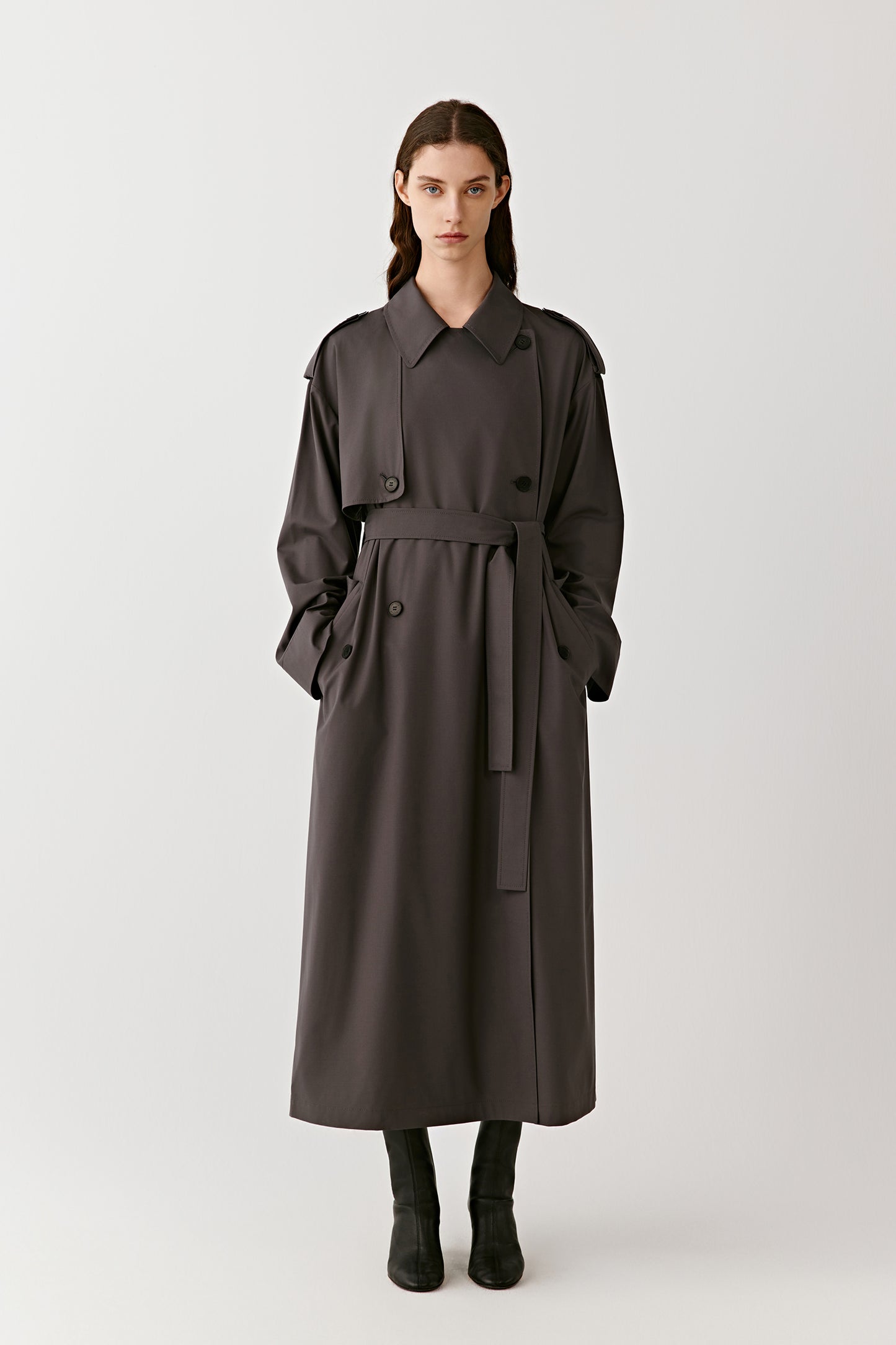 A woman stands wearing a grey double-breasted trench coat in wool and silk, the soft fabric flowing naturally around her.