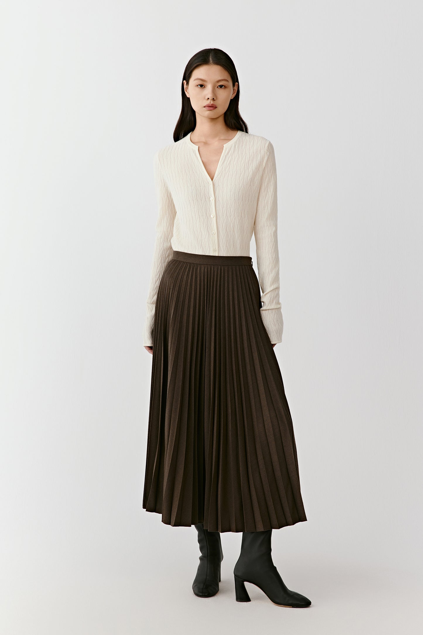 A woman wears a dark brown pleated wool flannel skirt with a light knit top, standing under even lighting.
