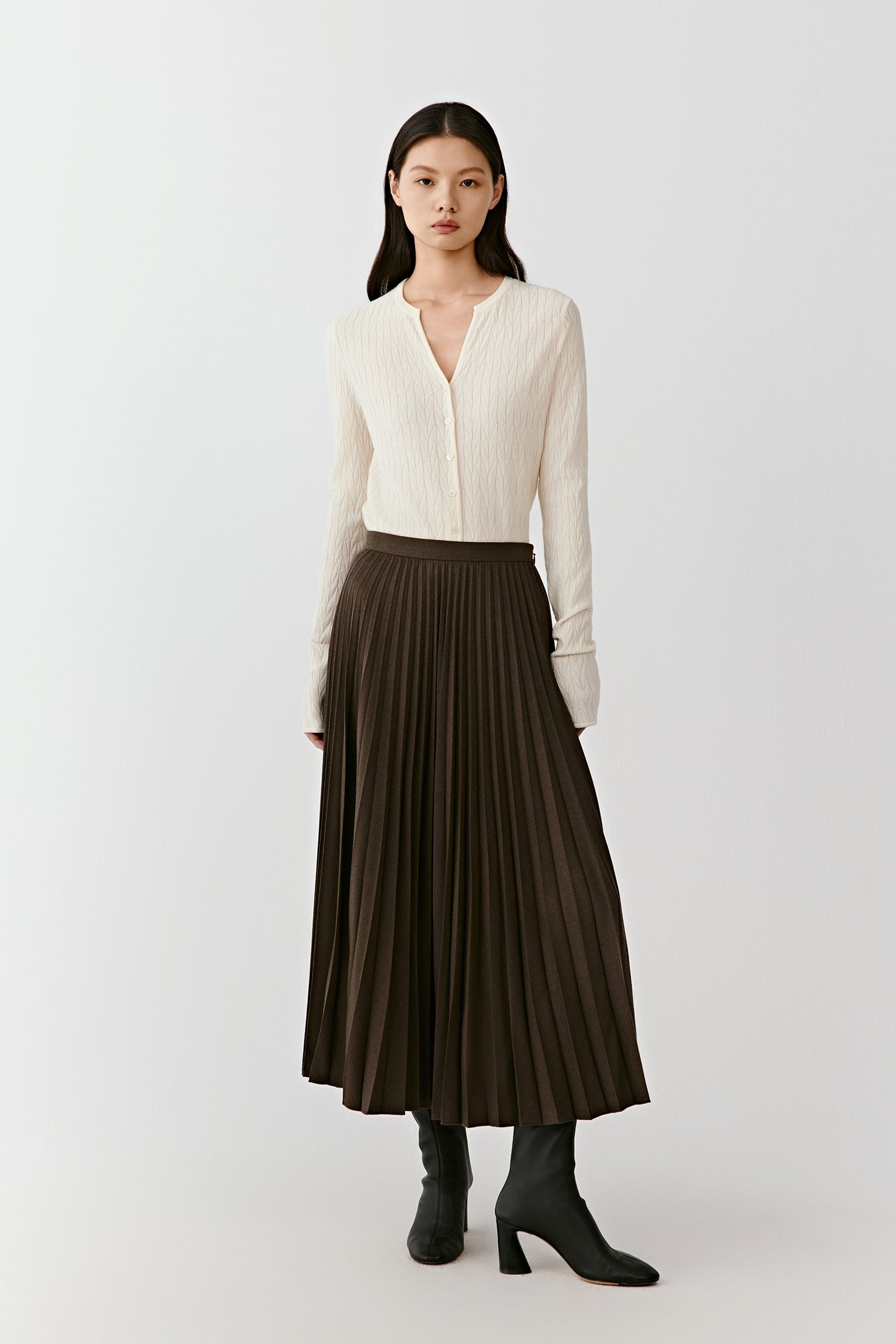 A woman wears a dark brown pleated wool flannel skirt with a light knit top, standing under even lighting.