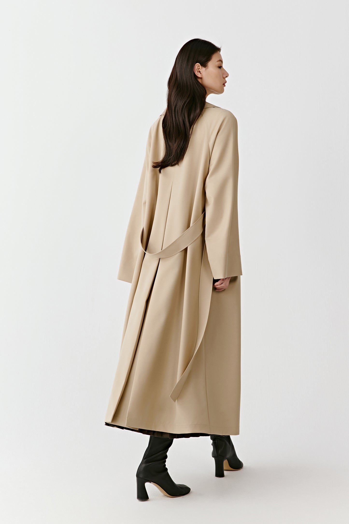 Back view of a woman wearing a long beige trench coat with a belt and pleated detail, standing on a white background.