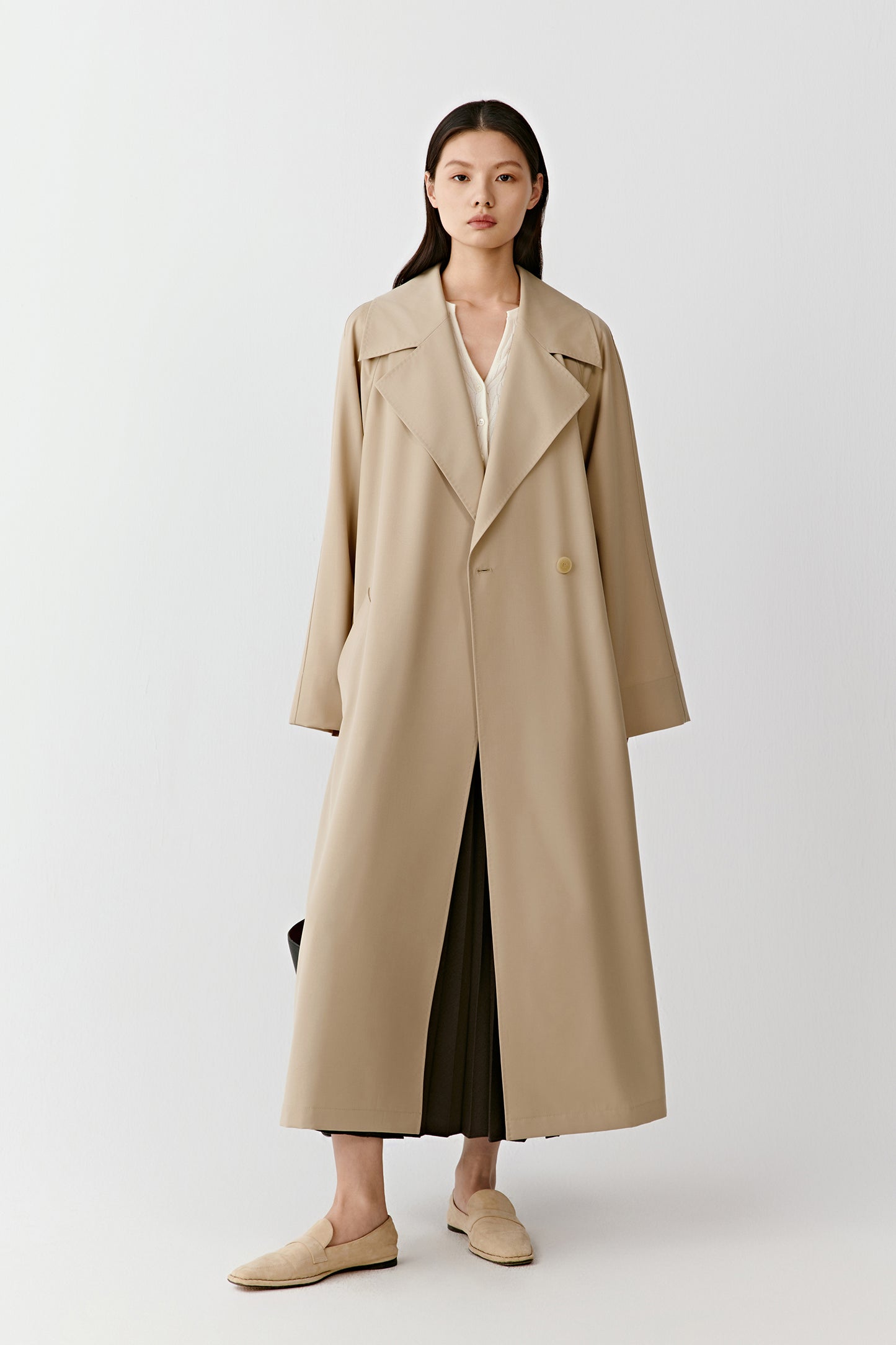 Woman wearing a long beige trench coat with wide lapels and side pockets, standing on a white background.