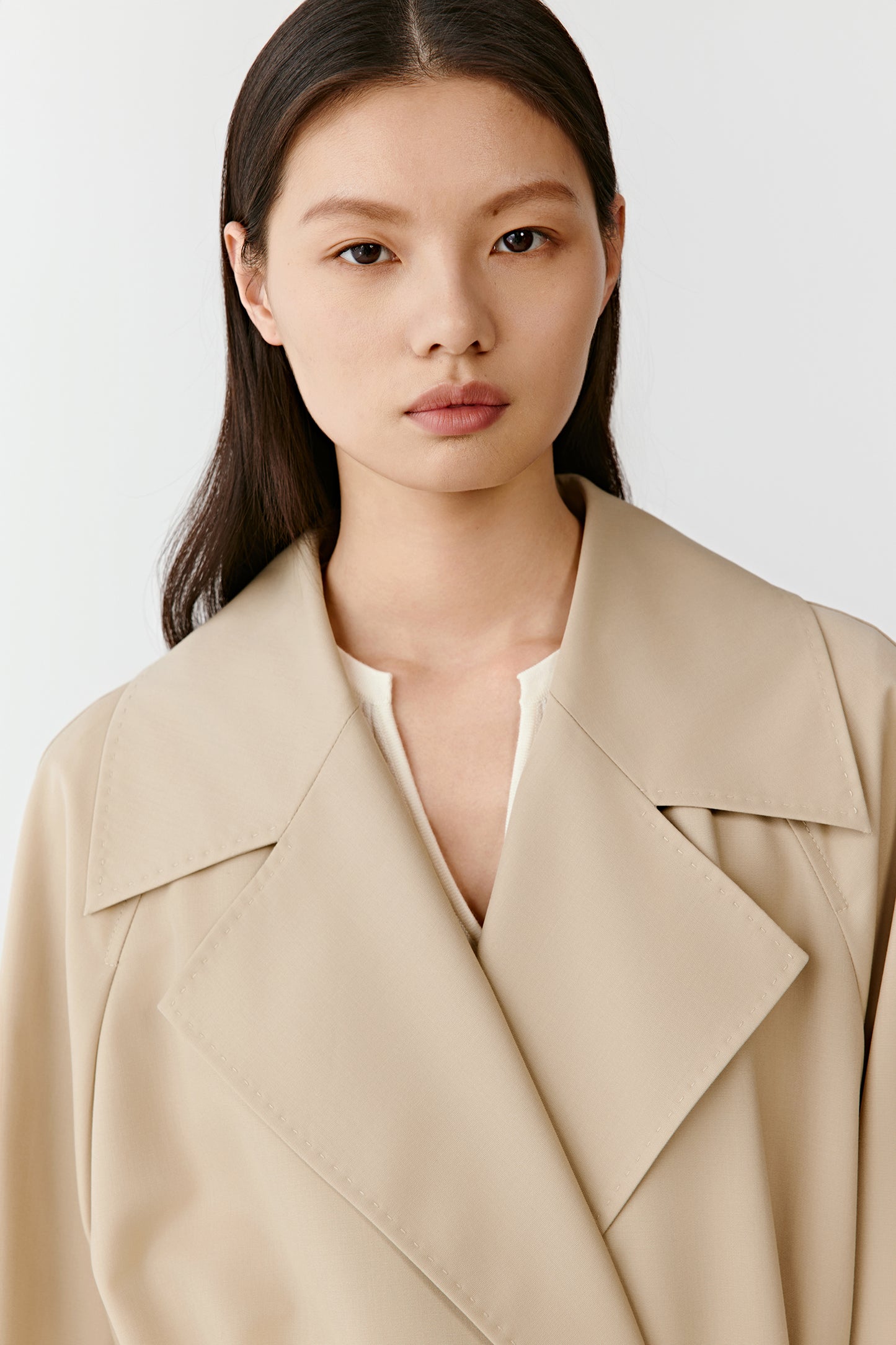 Close-up of a woman wearing a beige trench coat with wide lapels and a white inner top, on a white background.