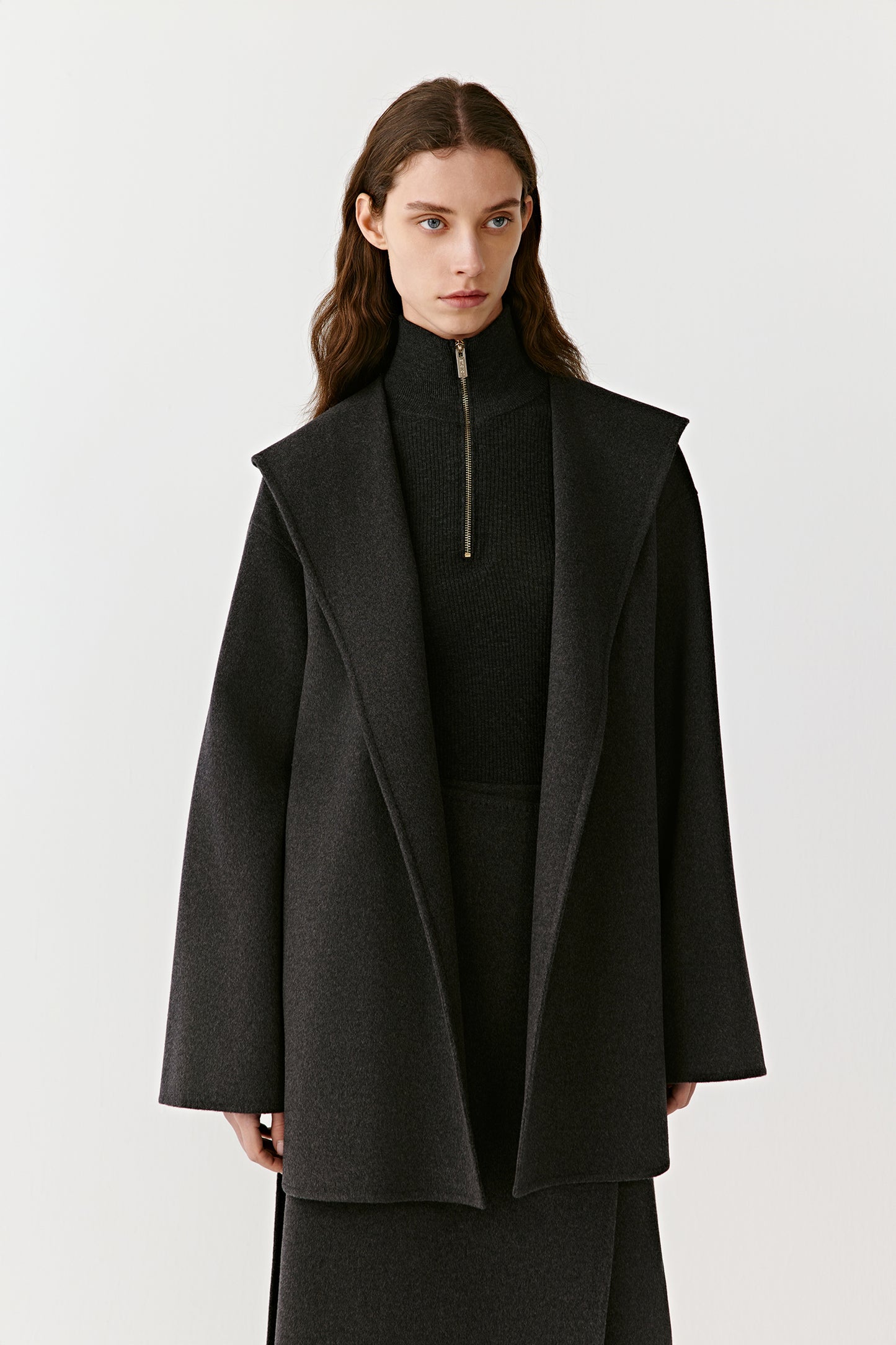 Woman wearing a long black wool coat with a hood and belted waist on a light background