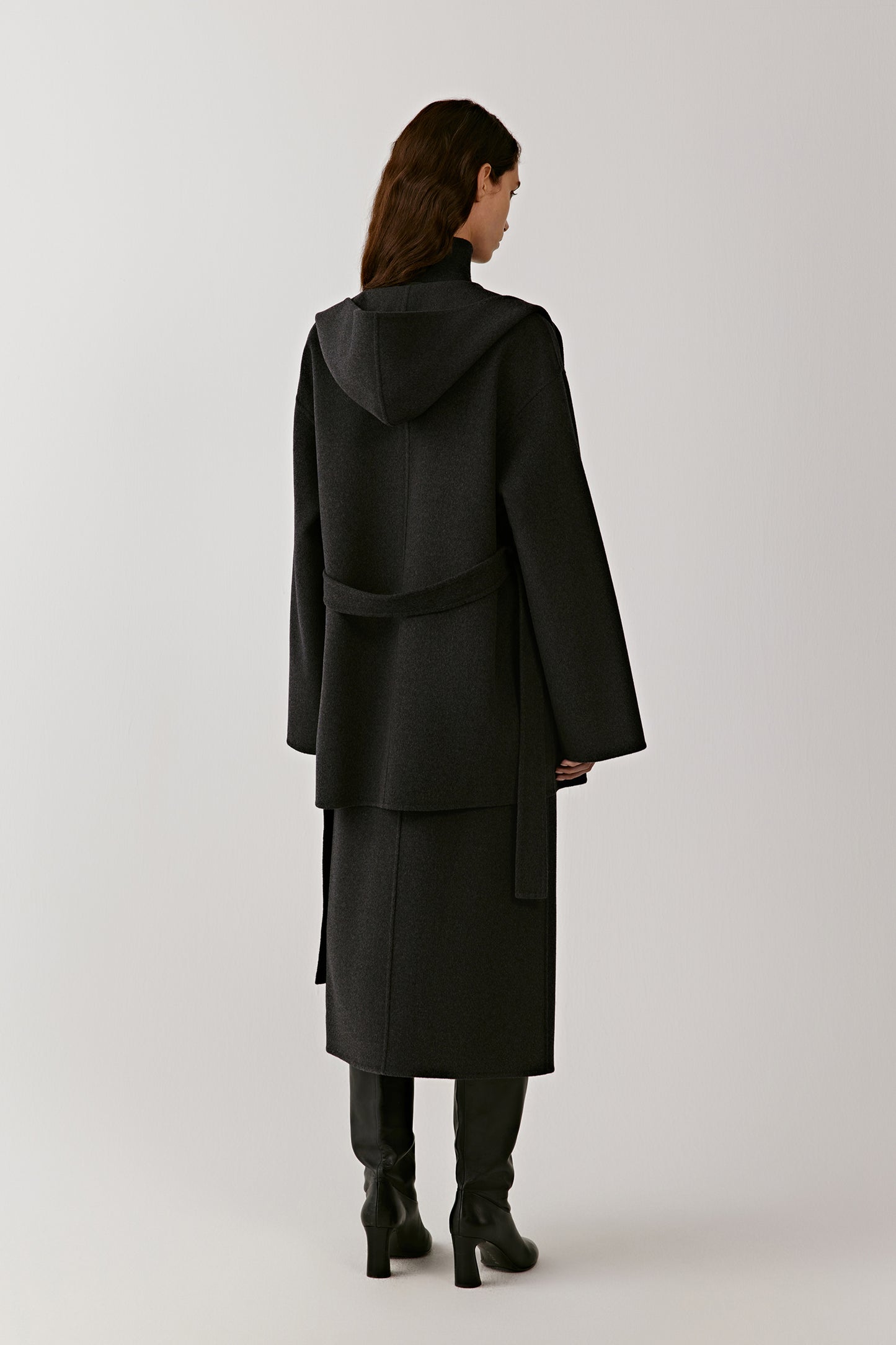 Back view of a long black hooded wool coat with belted waist on a light background