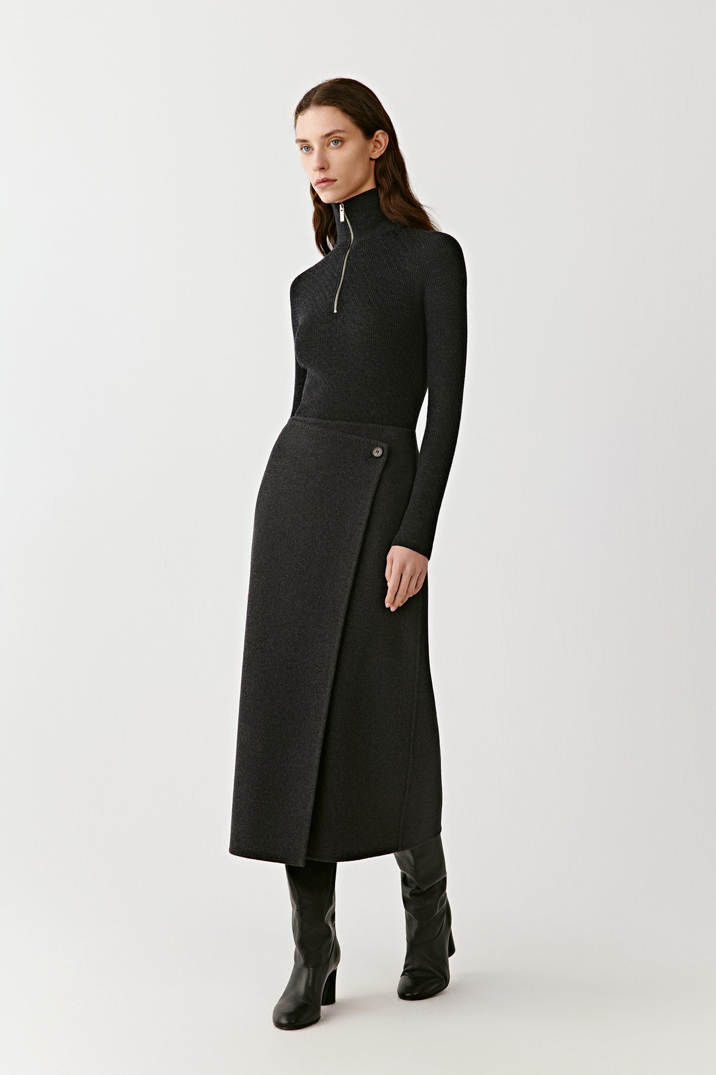 Model wearing a dark grey wrap midi skirt with a high waist and a fitted black turtleneck, paired with black boots. Minimal background, full-body front view.