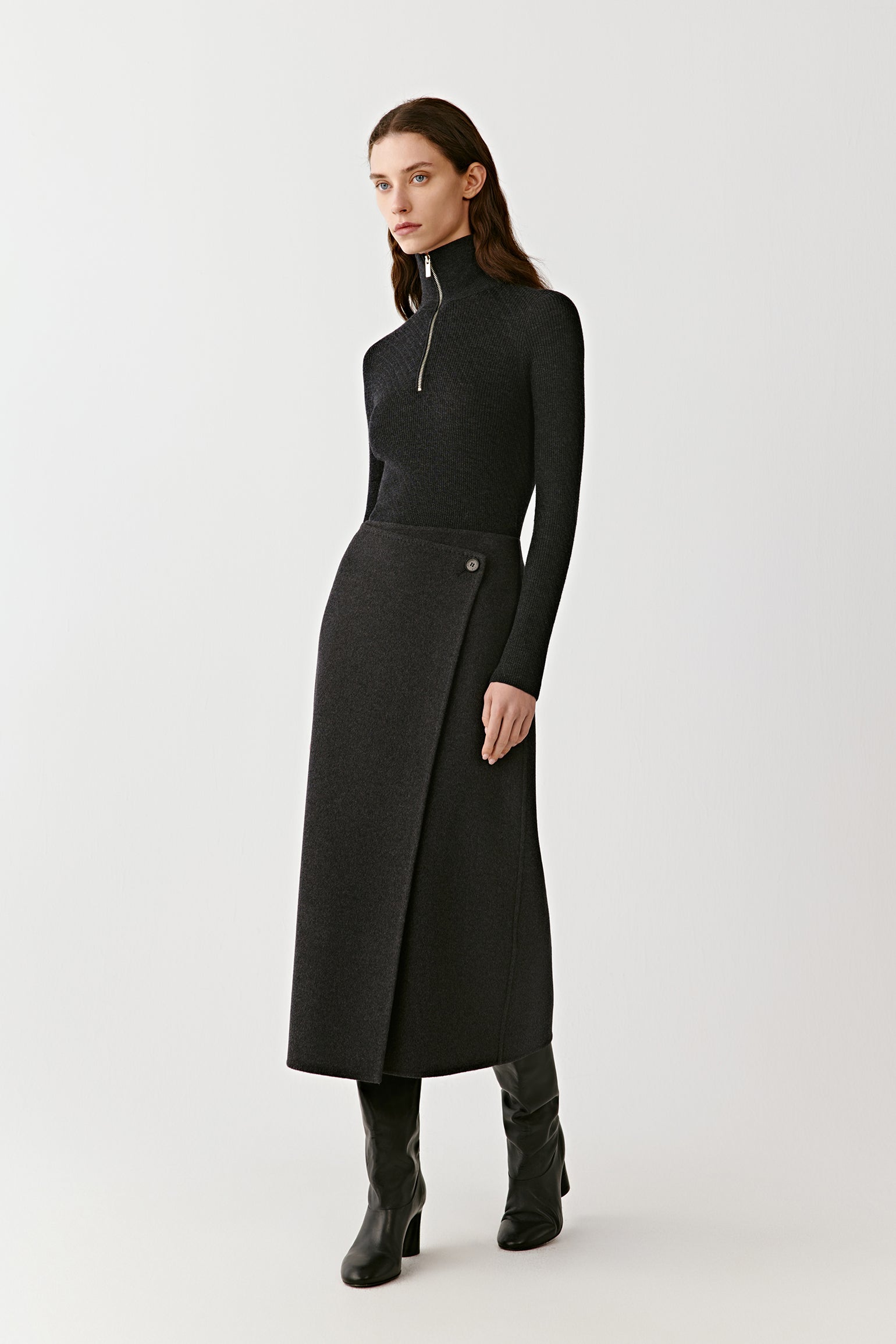 Model wearing a dark grey wrap midi skirt with a high waist and a fitted black turtleneck, paired with black boots. Minimal background, full-body front view.