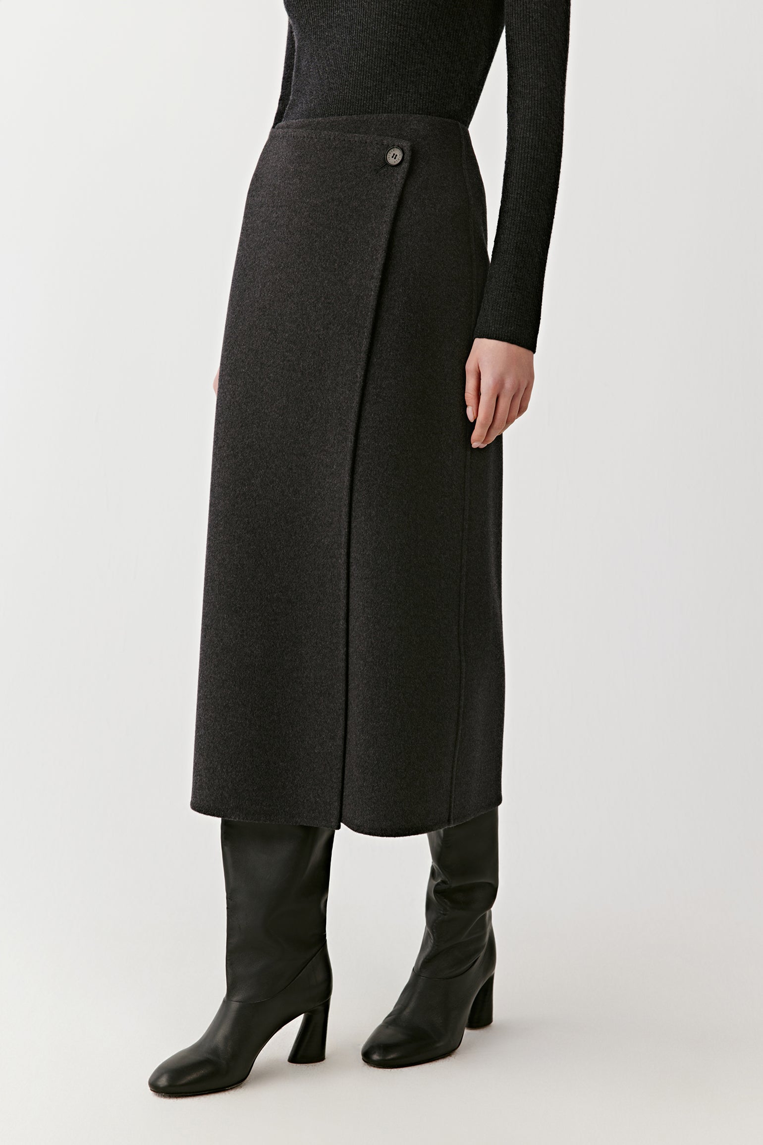 Side view of the model wearing a dark grey wrap skirt falling below the knee, paired with black high boots and a dark grey turtleneck.