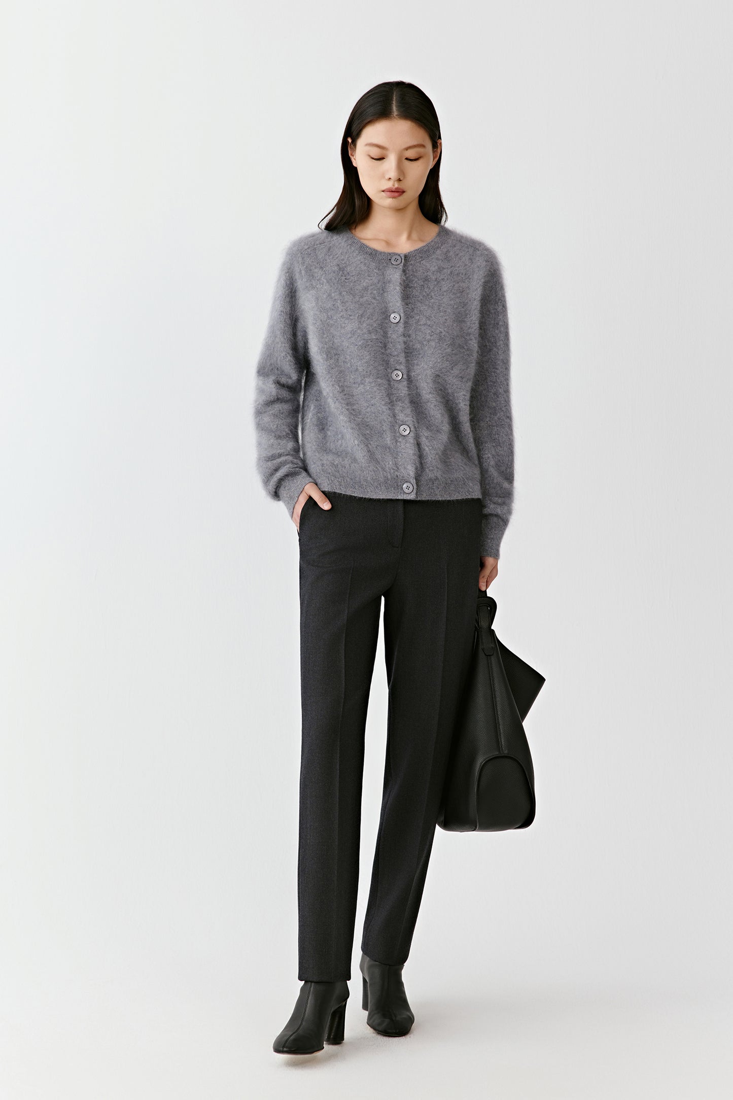 Model wearing a light gray crew-neck sweater tucked into black straight-leg tailored trousers with black heels and a black handbag.