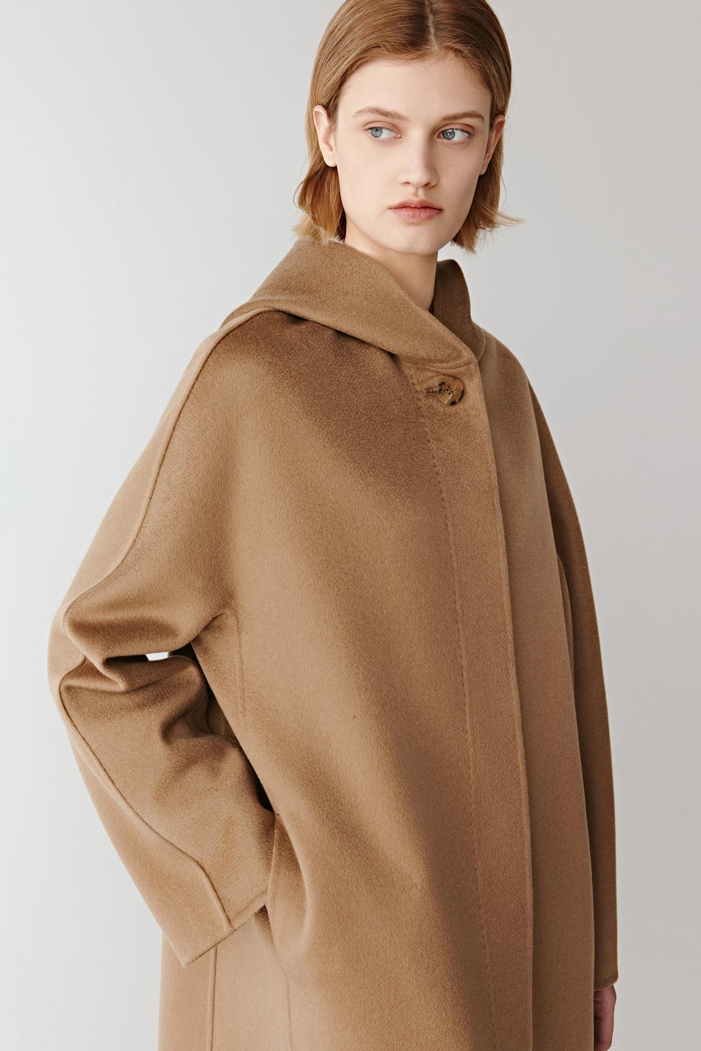 Model slightly turned to the side with hands in pockets, wearing a camel wool coat and matching pants.