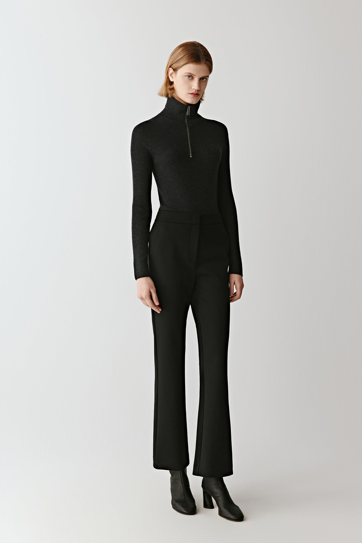 Full-length front view of the model wearing the deep black flared trousers in stretch combed wool, paired with a black turtleneck, showing the sleek and elongated silhouette.
