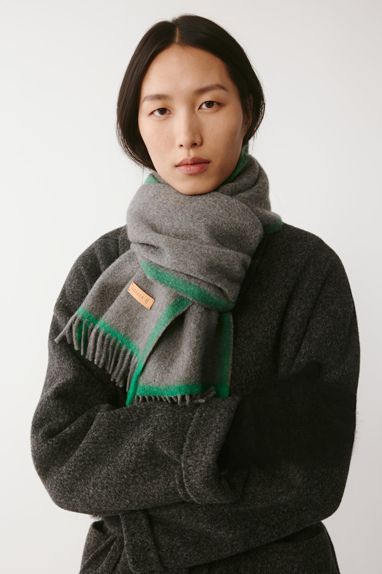 Woman wearing a grey wool scarf with green trim styled over a dark textured coat on a light background