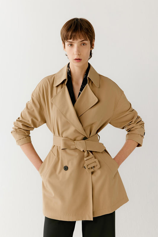 Three-quarter view showing the full silhouette and belted construction of the trench coat. The image emphasizes the fluid drape of the fabric, the softly structured shape and the clean lines that define the garment. The camel shade enhances the subtle depth and refined character of the design.