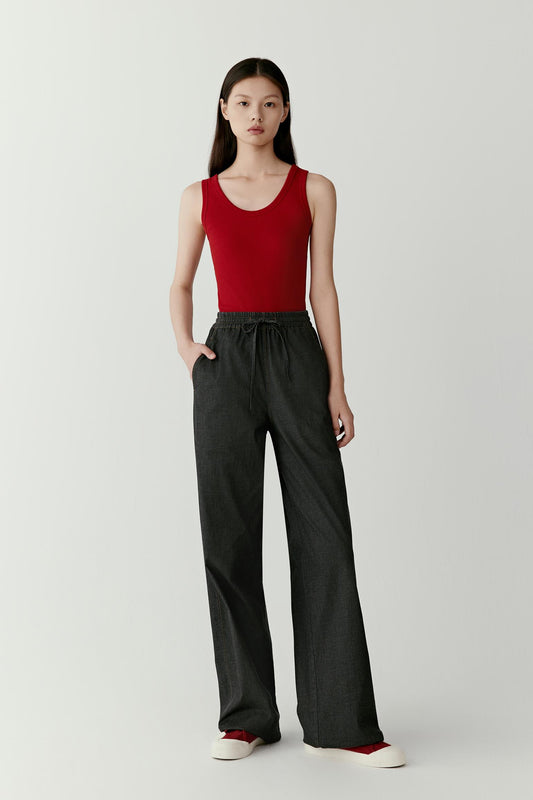 Full-length front view showing the tank top styled with dark wide-leg trousers. The fitted shape follows the body closely, while the lightweight rib jersey provides flexibility and comfort. The simple construction creates a streamlined silhouette with clear proportions.