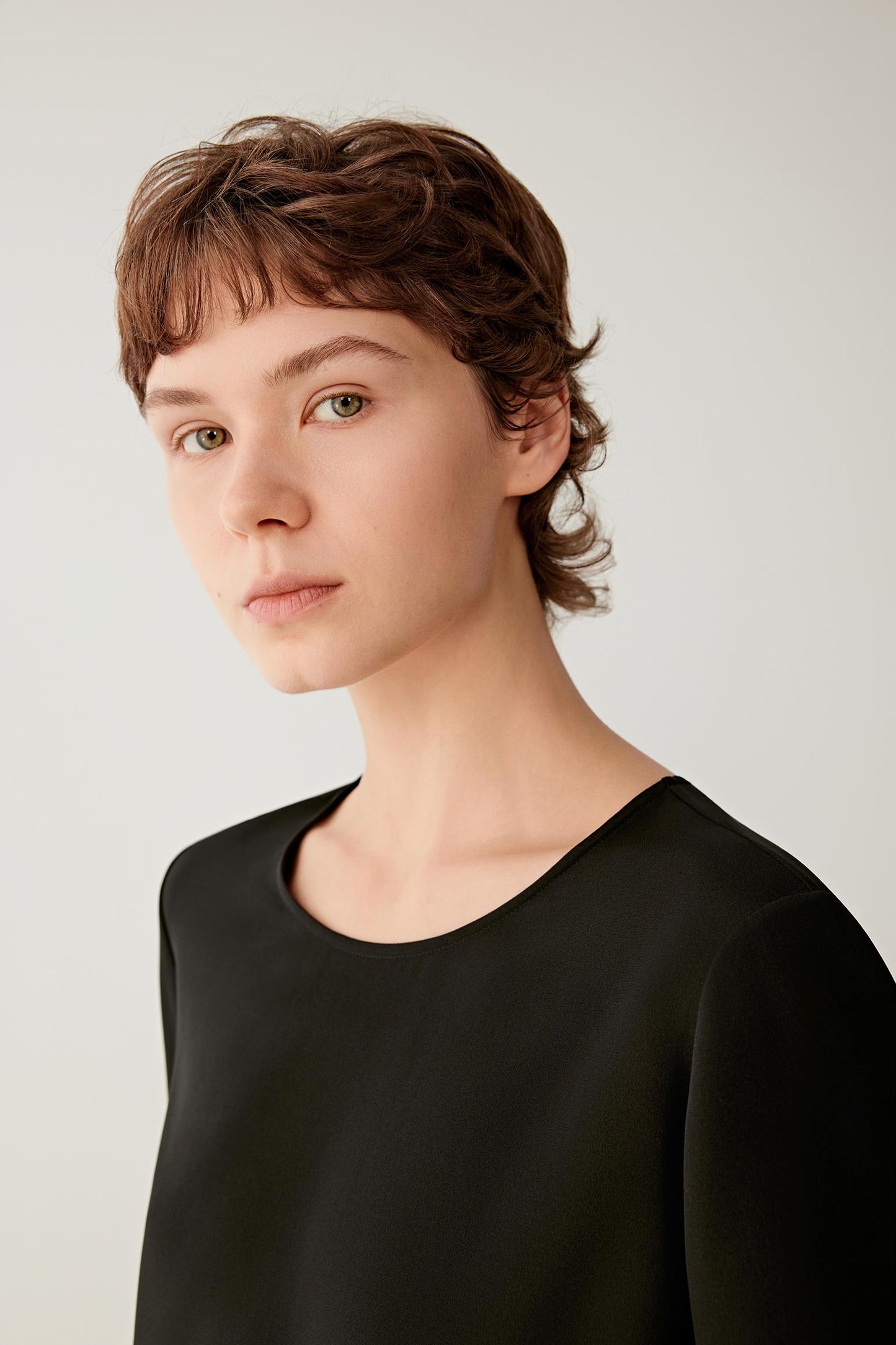 Close-up portrait focusing on the wide round neckline, shoulder line and upper section of the garment. The image reveals the smooth texture, soft finish and subtle lustre characteristic of silk crêpe de Chine, while emphasizing the clean construction and minimalist design.