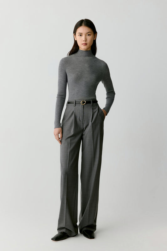 A woman stands against a neutral backdrop wearing a smoke-grey ribbed pullover paired with tailored trousers. The composition highlights the sleek, tonal palette.