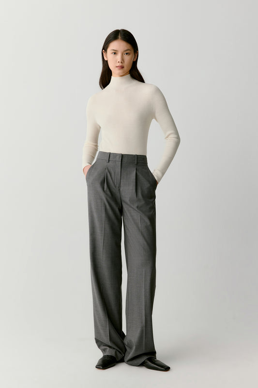 A woman stands facing the camera wearing an ivory ribbed pullover in superfine wool, tucked into soft grey trousers. The light background enhances the purity of the tones.