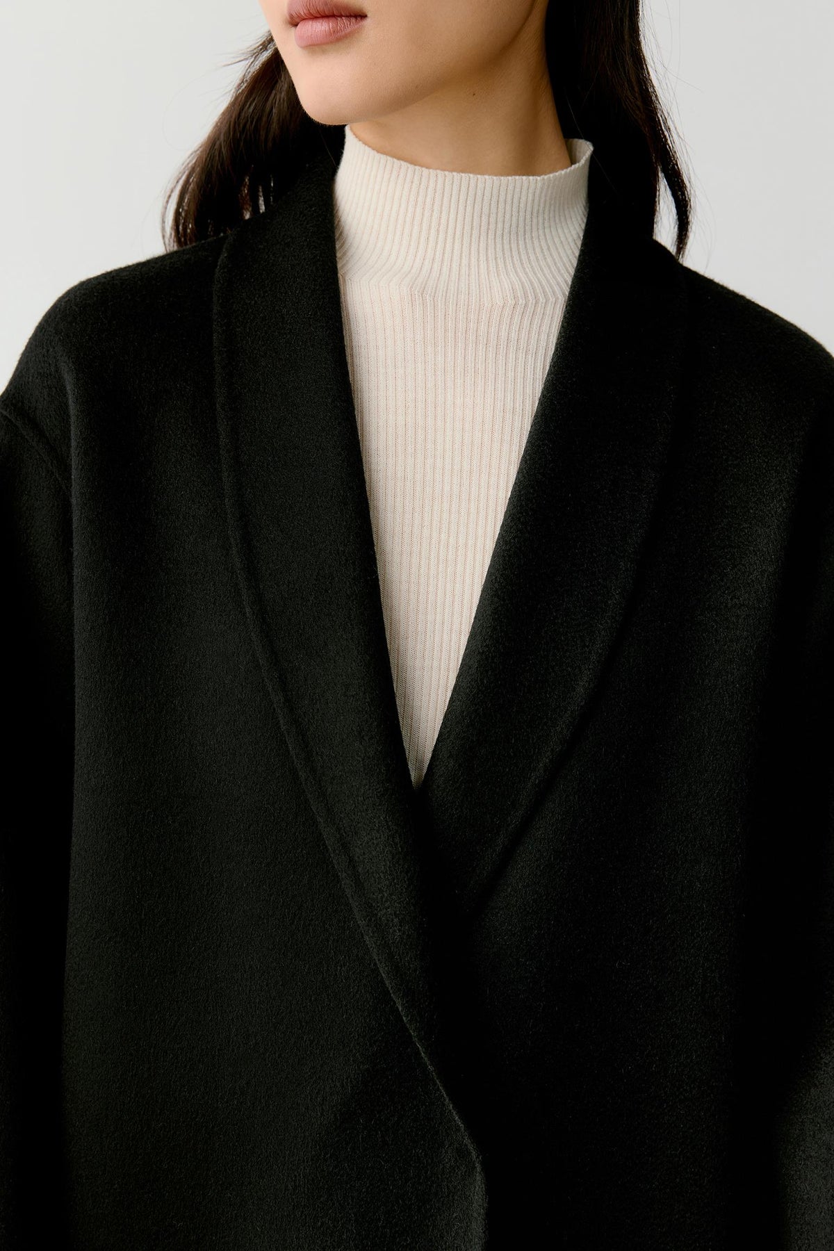 A close-up of a model wearing a black shawl collar wrap coat over a cream ribbed turtleneck sweater, set against a plain light gray background.