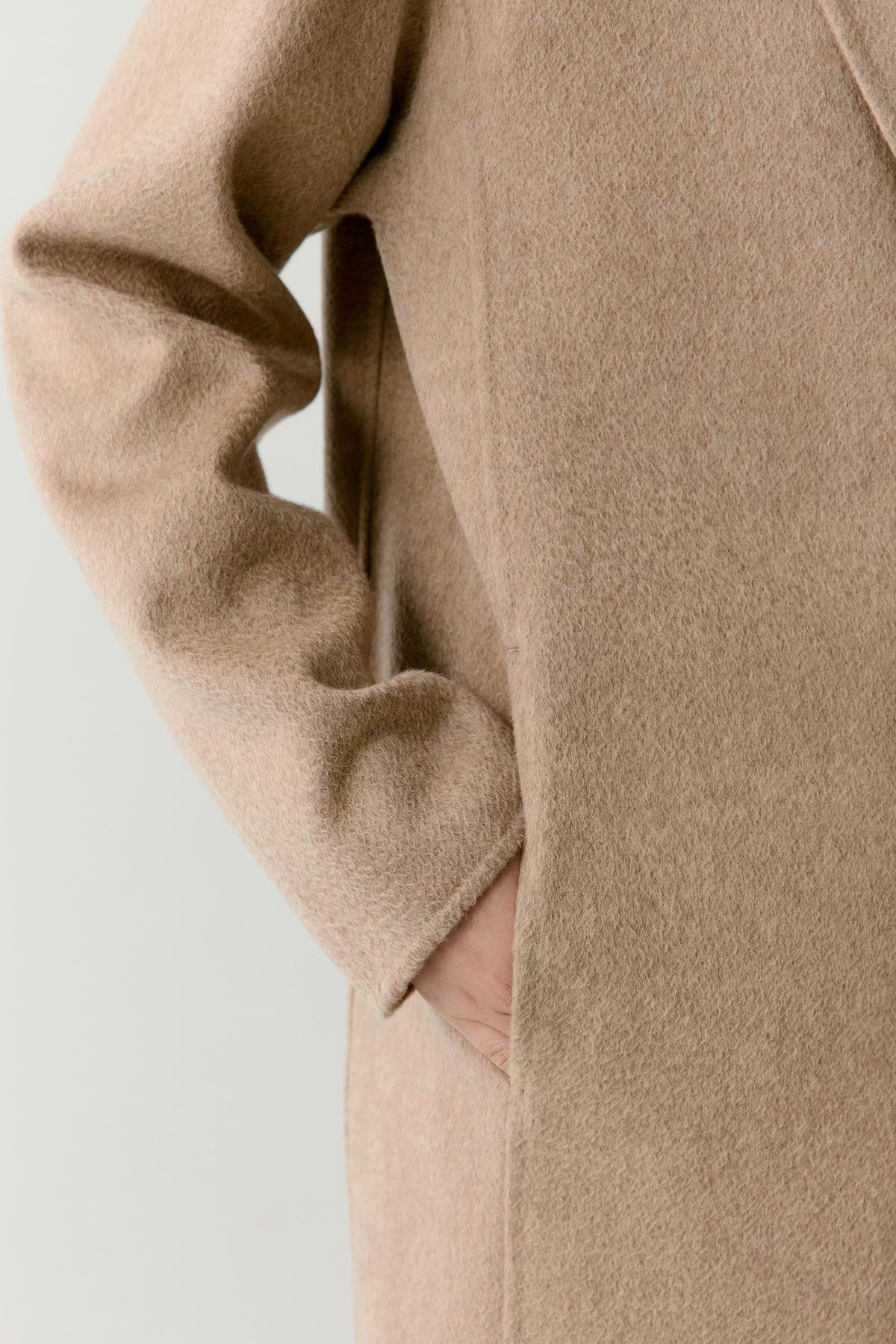 Close-up of a person wearing a soft camel-colored wool coat, showing the sleeve and side pocket with a hand partially visible.